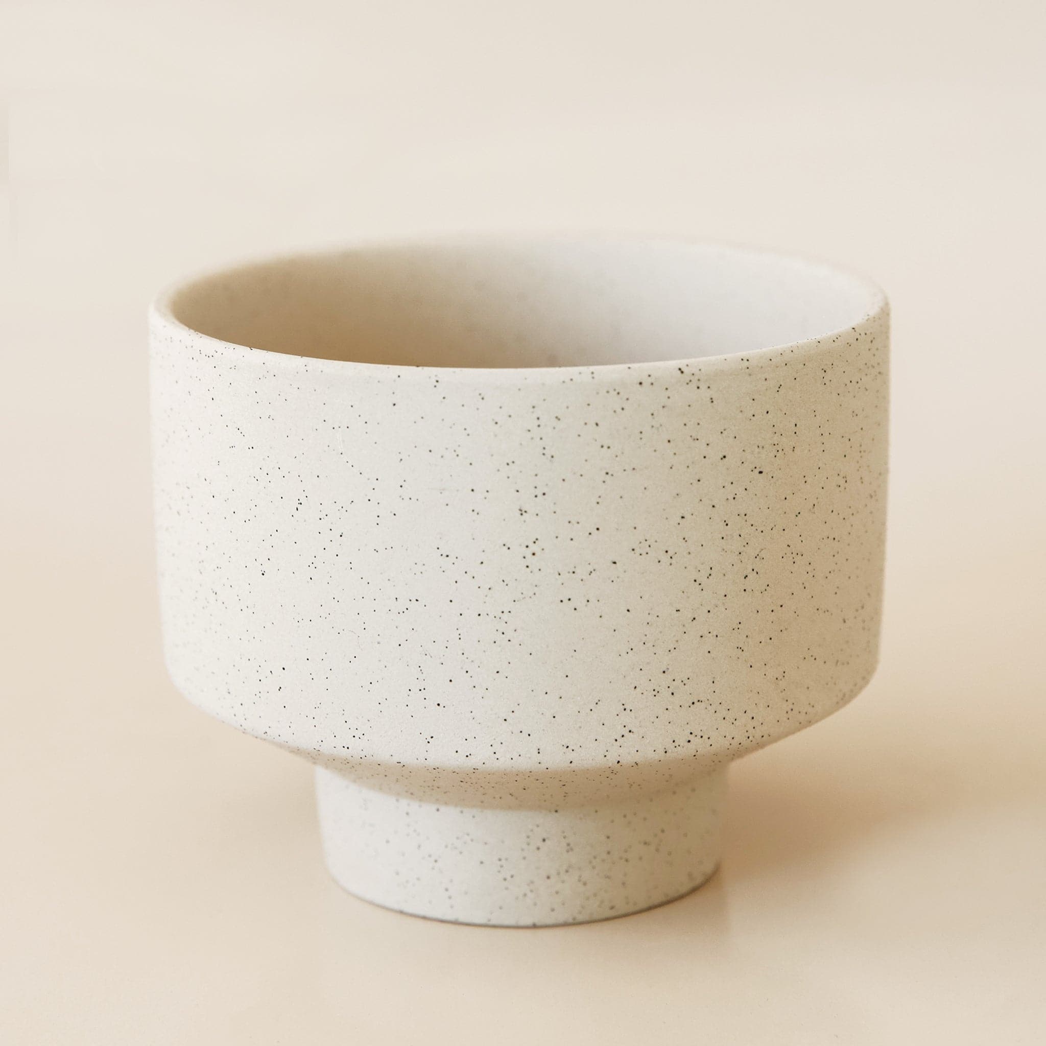 In front of a tan background is a round, ceramic dove pot with a tapered bottom. The pot has black speckles.