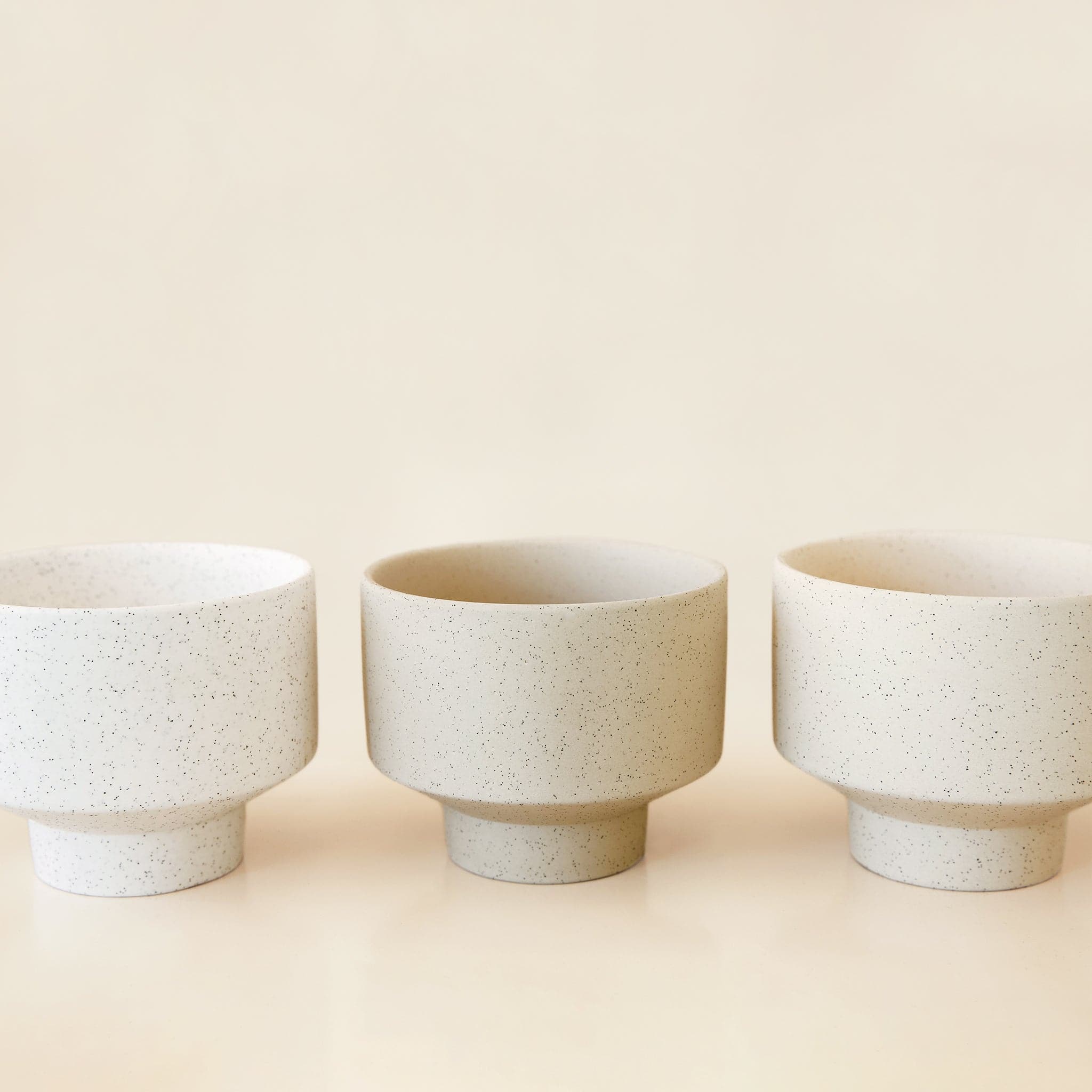 Three round, ceramic white pots with a tapered bottom. The pots have black speckles and are in an array of warm cream tones. 