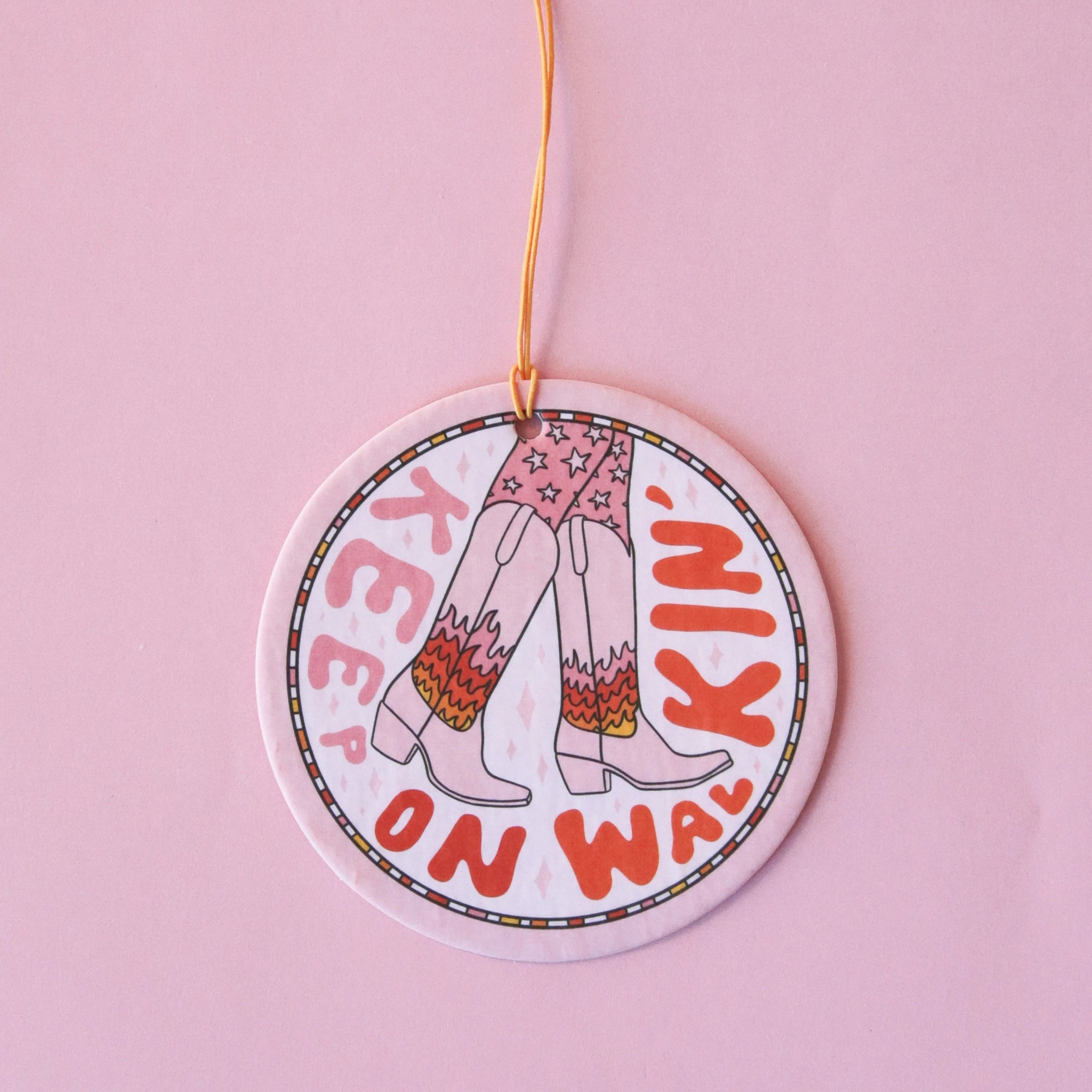 A light pink circle air freshener with a elastic loop for hanging and a cowgirl boot graphic with fire details on them and text that reads, "Keep On Walking'" in keep and red text around the edge of the air freshener.