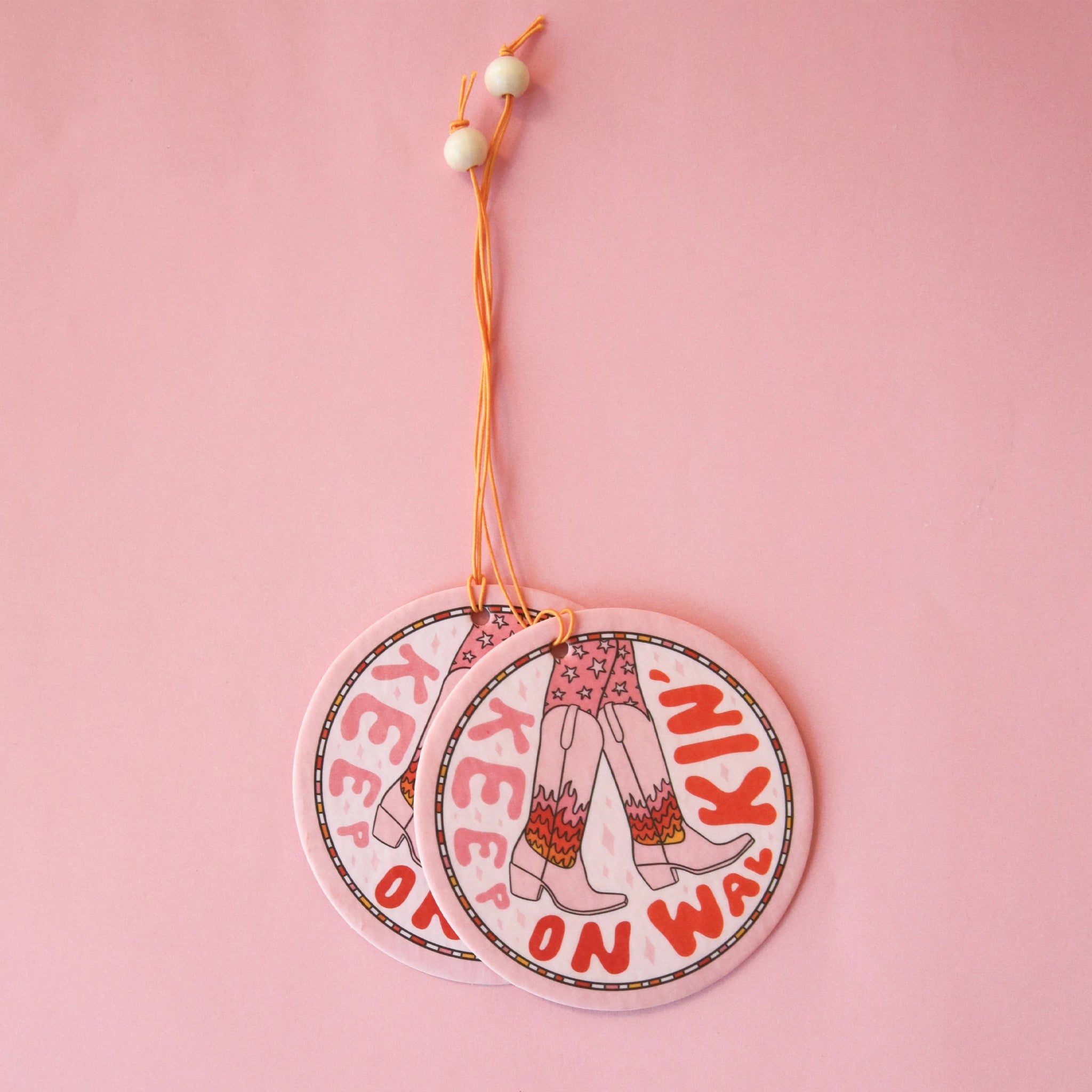 A light pink circle air freshener with a elastic loop for hanging and a cowgirl boot graphic with fire details on them and text that reads, "Keep On Walking'" in keep and red text around the edge of the air freshener.