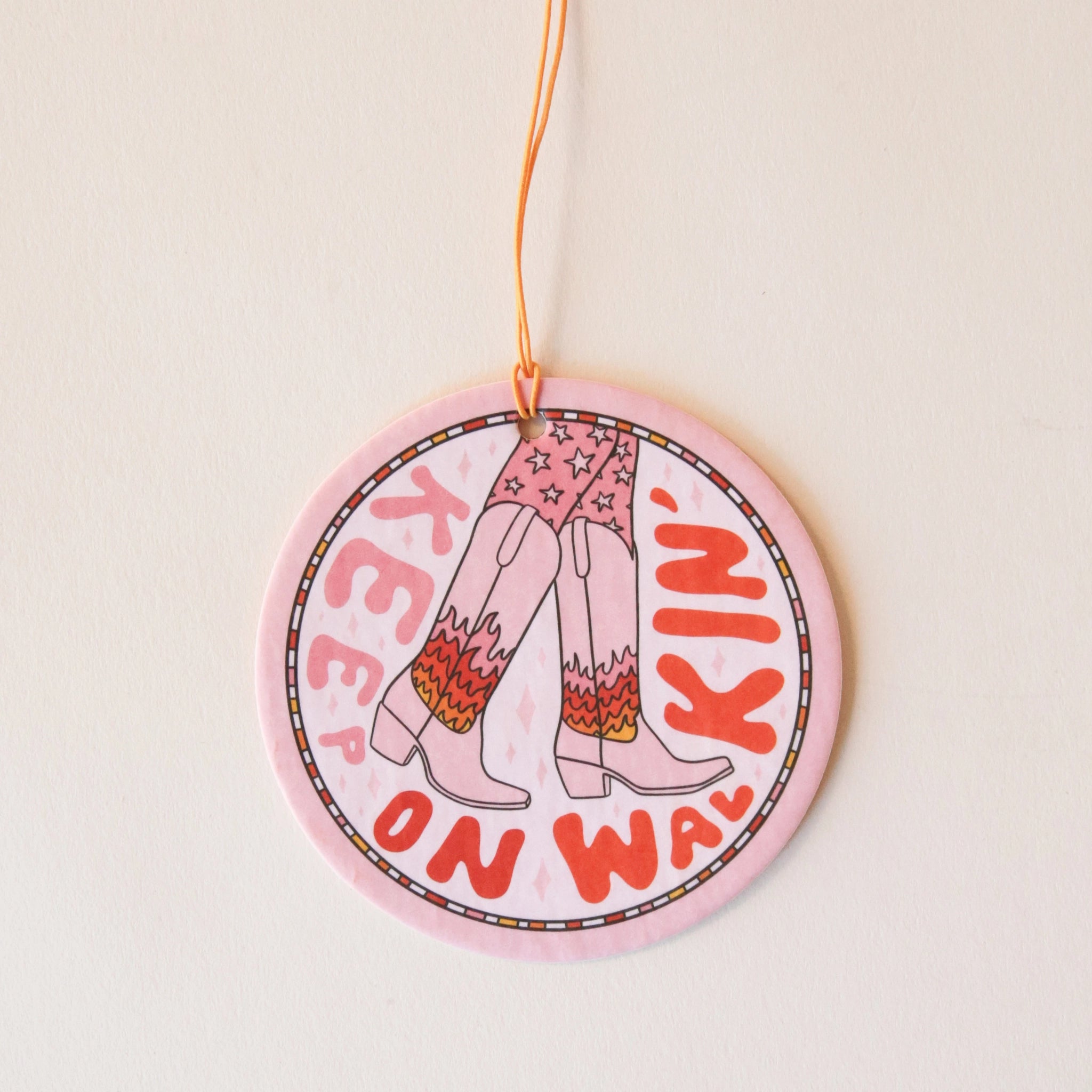 A light pink circle air freshener with a elastic loop for hanging and a cowgirl boot graphic with fire details on them and text that reads, &quot;Keep On Walking&#39;&quot; in keep and red text around the edge of the air freshener. 