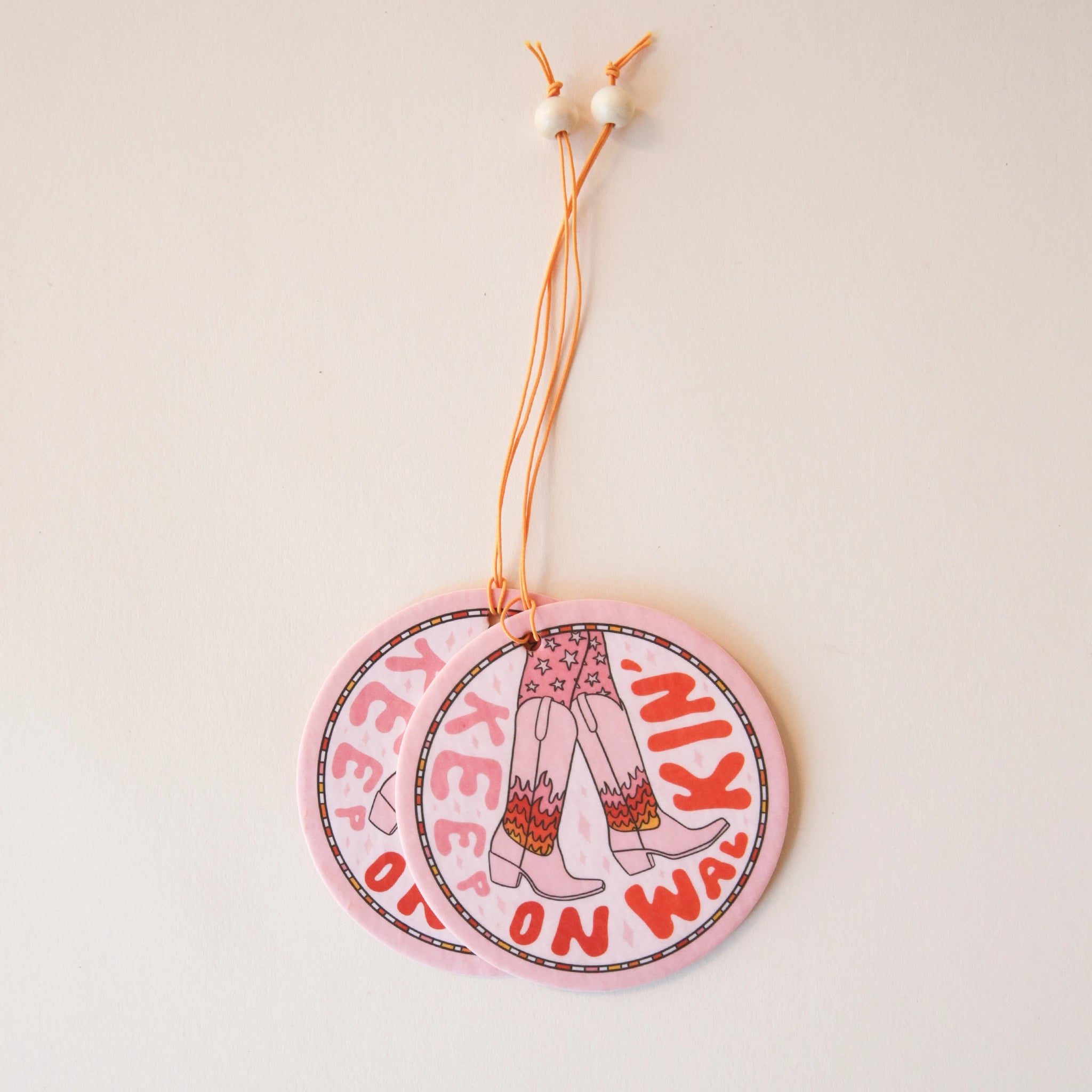 A light pink circle air freshener with a elastic loop for hanging and a cowgirl boot graphic with fire details on them and text that reads, &quot;Keep On Walking&#39;&quot; in keep and red text around the edge of the air freshener.
