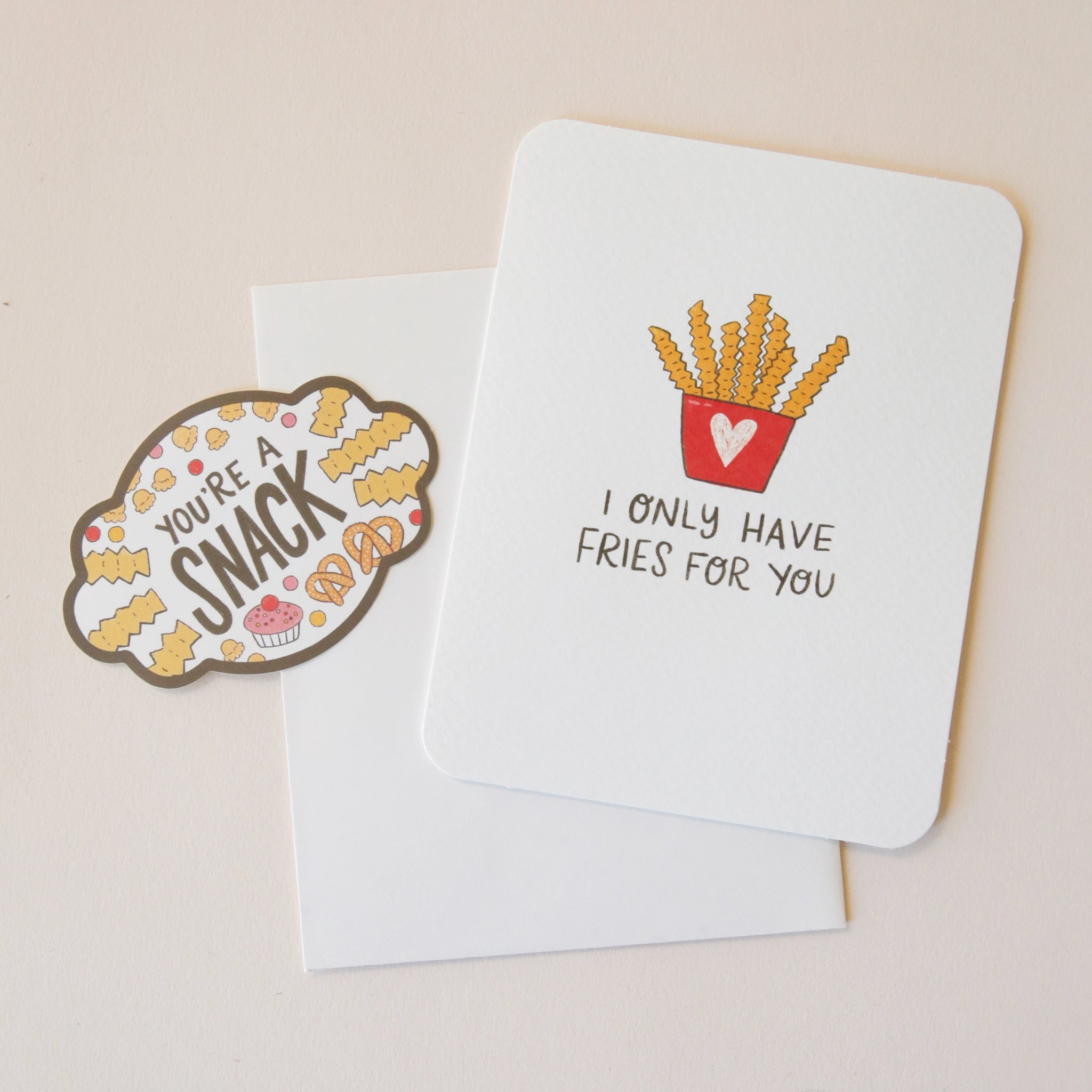A white card with an illustration of a red carton with a pink heart on the front holding crinkle cut French fries and black text that reads, "I Only Have Fries For You" as well as a coordinating sticker that says, "You're A Snack" along with a border of fries, popcorn, pretzels and cupcakes.
