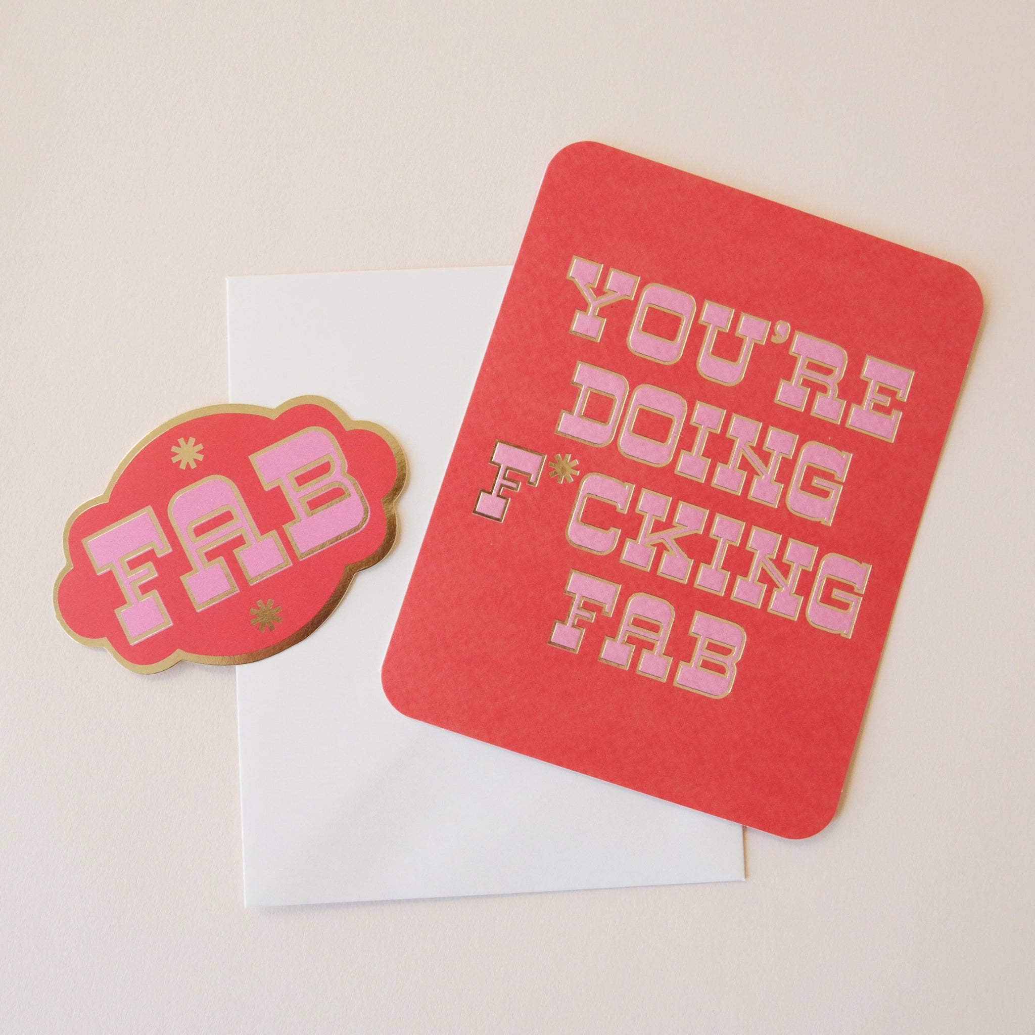 A red card with pink western style text that reads, "You're Doing F*cking Fab" along with a white envelope and a coordinating red sticker that has the same style font and says, "FAB".