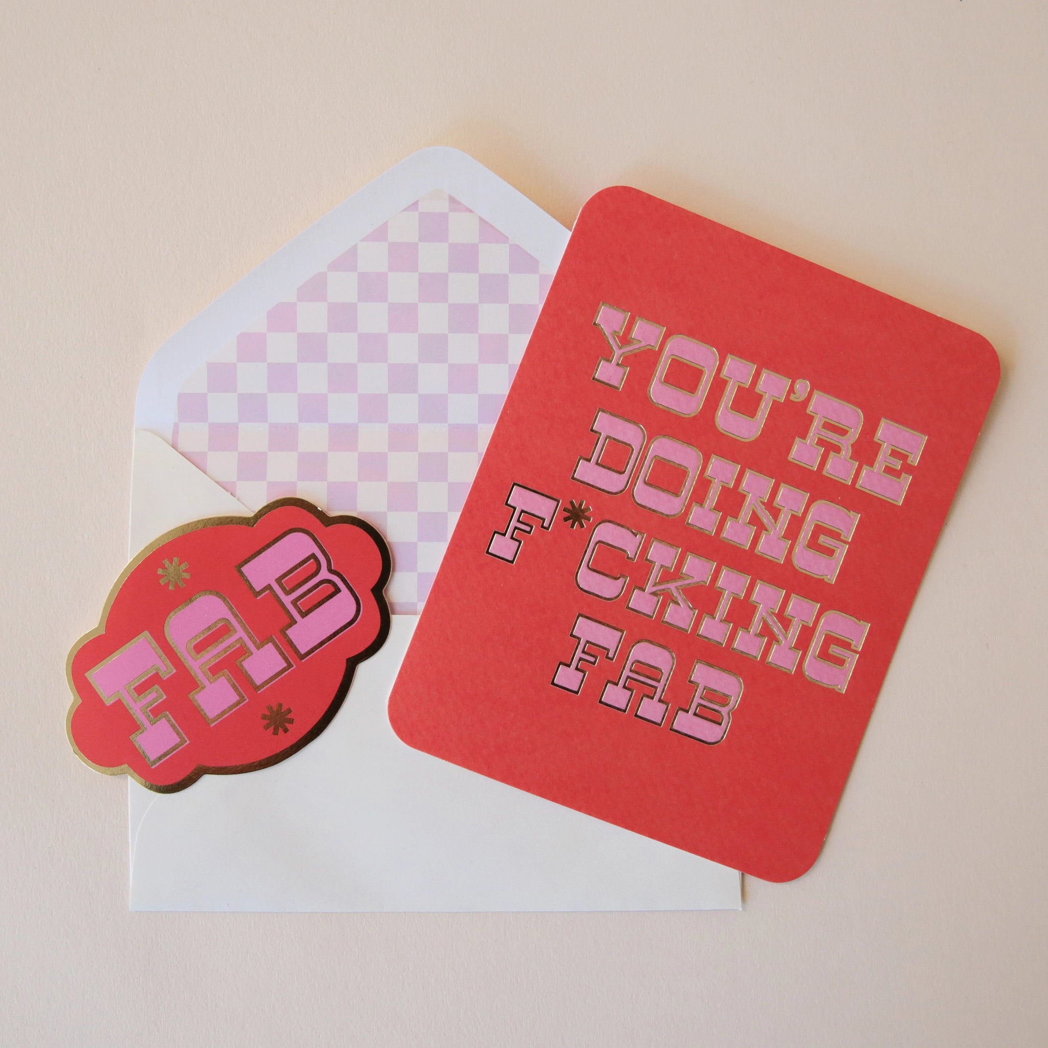 A red card with pink western style text that reads, "You're Doing F*cking Fab" along with a white envelope and a coordinating red sticker that has the same style font and says, "FAB". The interior of the white envelope has a light pink and white checker design.
