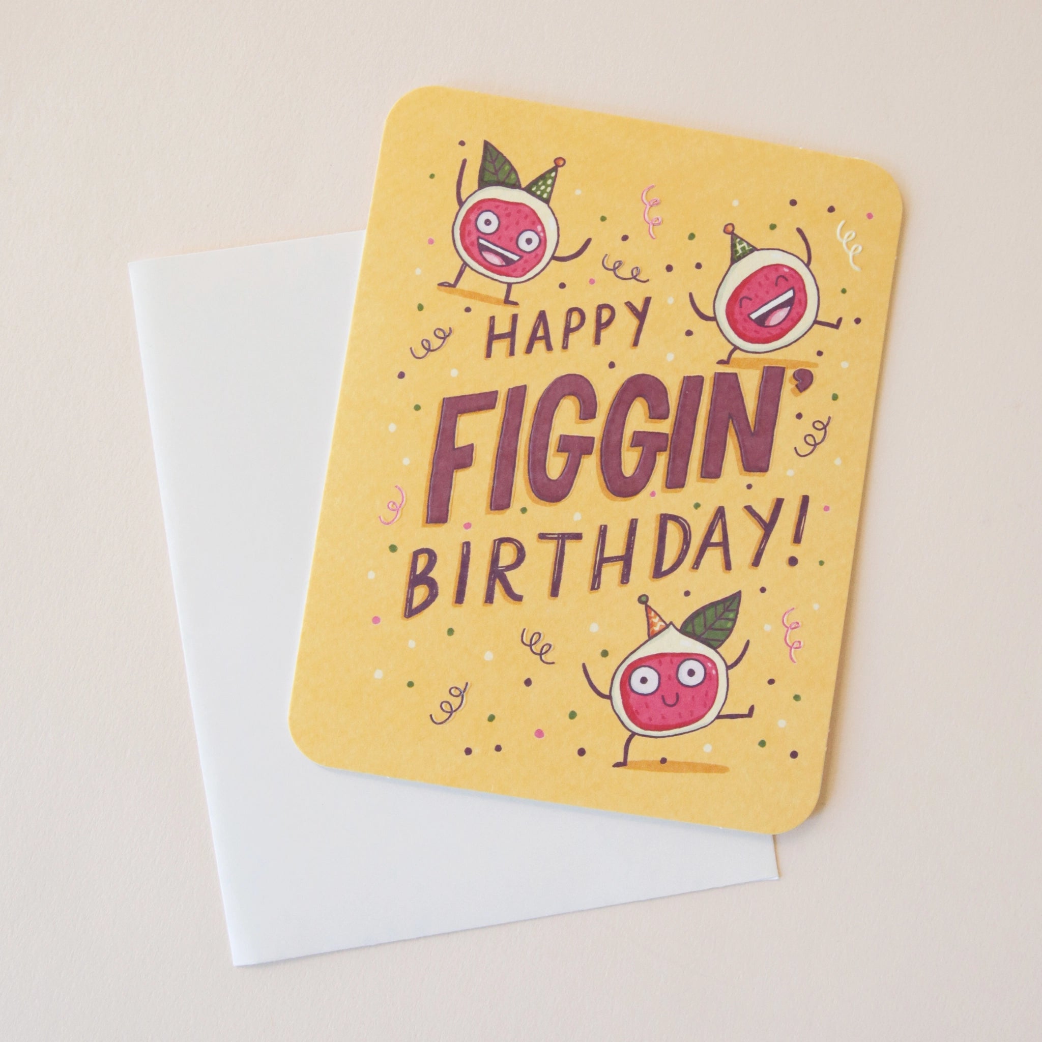 A yellow card with three fig illustrations with their hands up and celebrating along with words in the center that read, "Happy Figgin' Birthday" along with a coordinating sticker that reads, "Get Figgy With It" and a white envelope.
