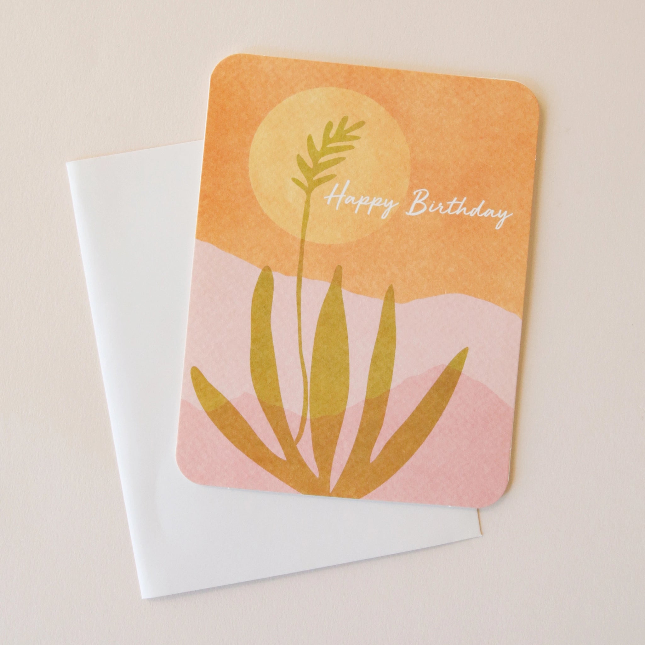 A card with a desert scape illustration with an aloe plant, an orange sky, a yellow sun and a pink mountainous background and white text that reads, "Happy Birthday". Also included is a sticker that says "Best Wishes" with the same illustration.