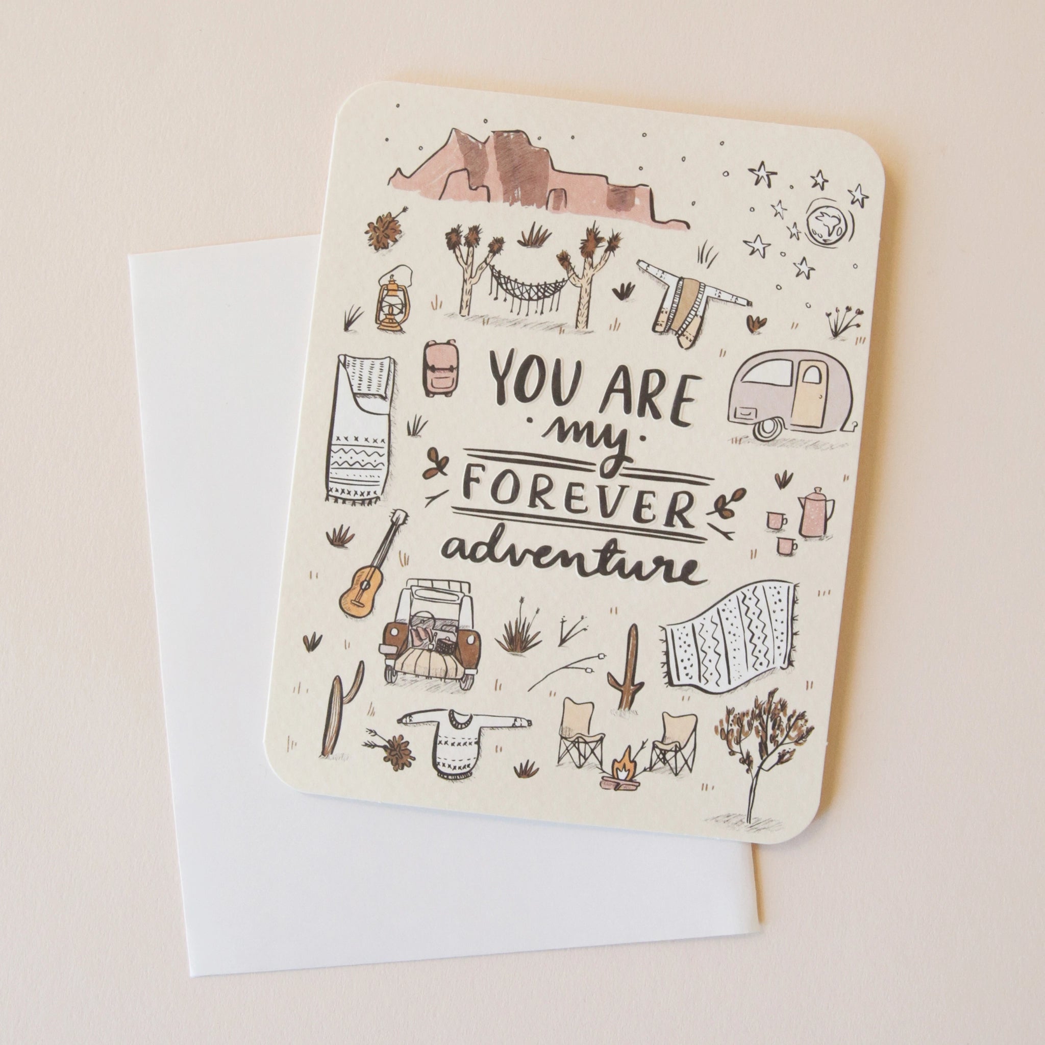 On a neutral background is an ivory card with text in the center that reads, "You Are My Forever Adventure" along with illustrations of various desert camping and road-tripping items, like a camper, a hammock, stars, a campfire and chairs etc as well as a sticker that is included with purchase.