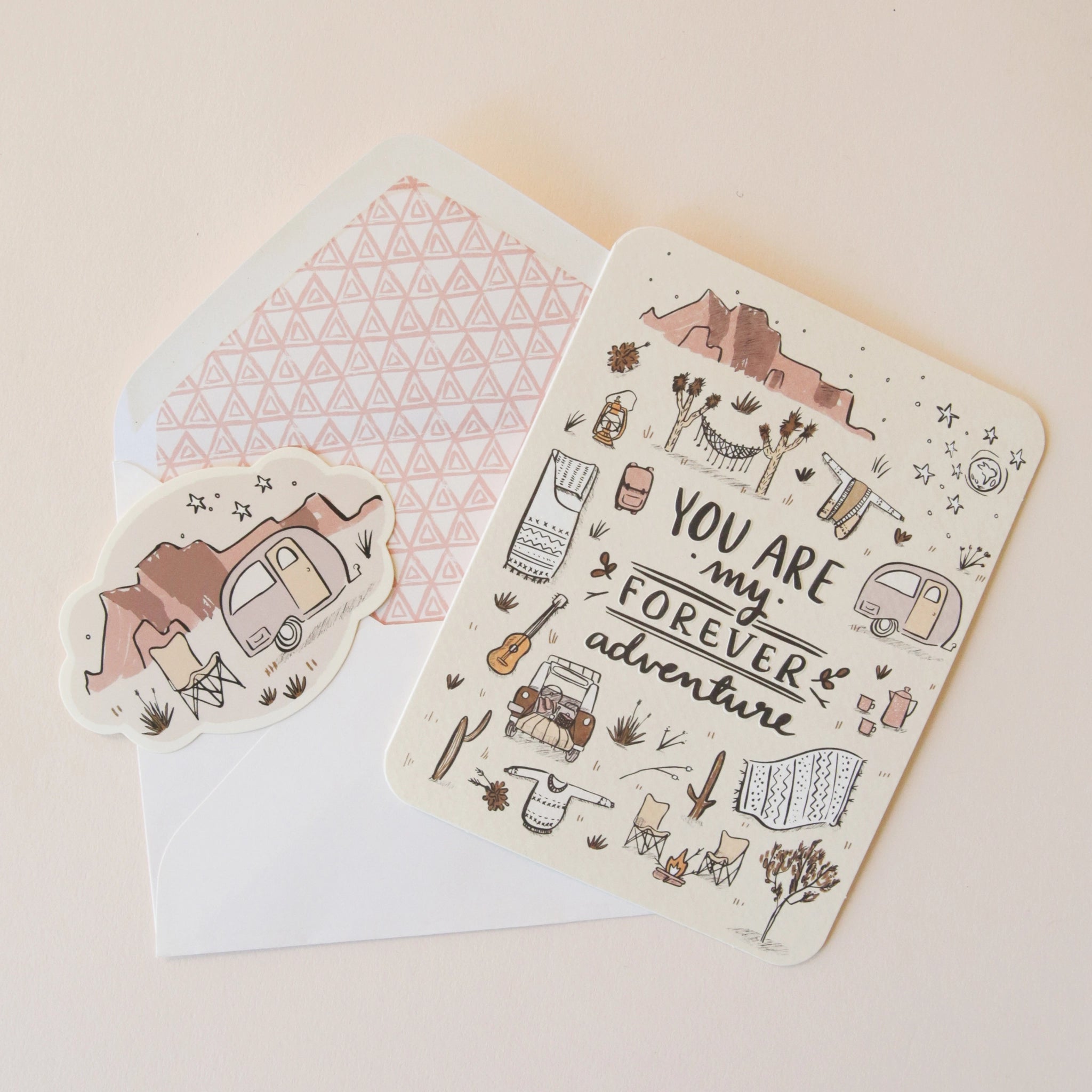 On a neutral background is an ivory card with text in the center that reads, "You Are My Forever Adventure" along with illustrations of various desert camping and road-tripping items, like a camper, a hammock, stars, a campfire and chairs etc as well as a sticker that is included with purchase.