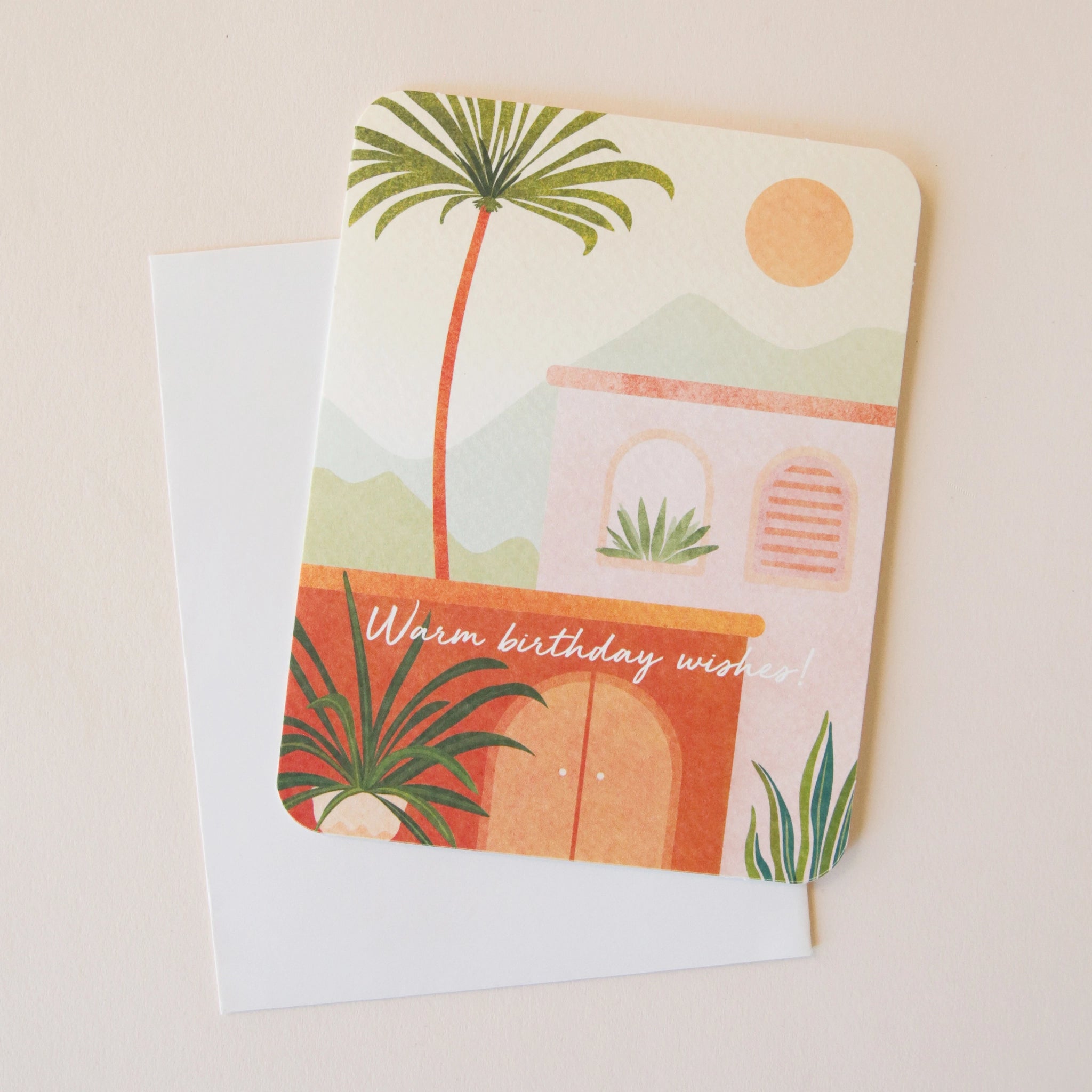 On a neutral background is a card with a sun and palm tree graphic along with a coordinating sticker and text along the front that reads, "Warm birthday wishes".
