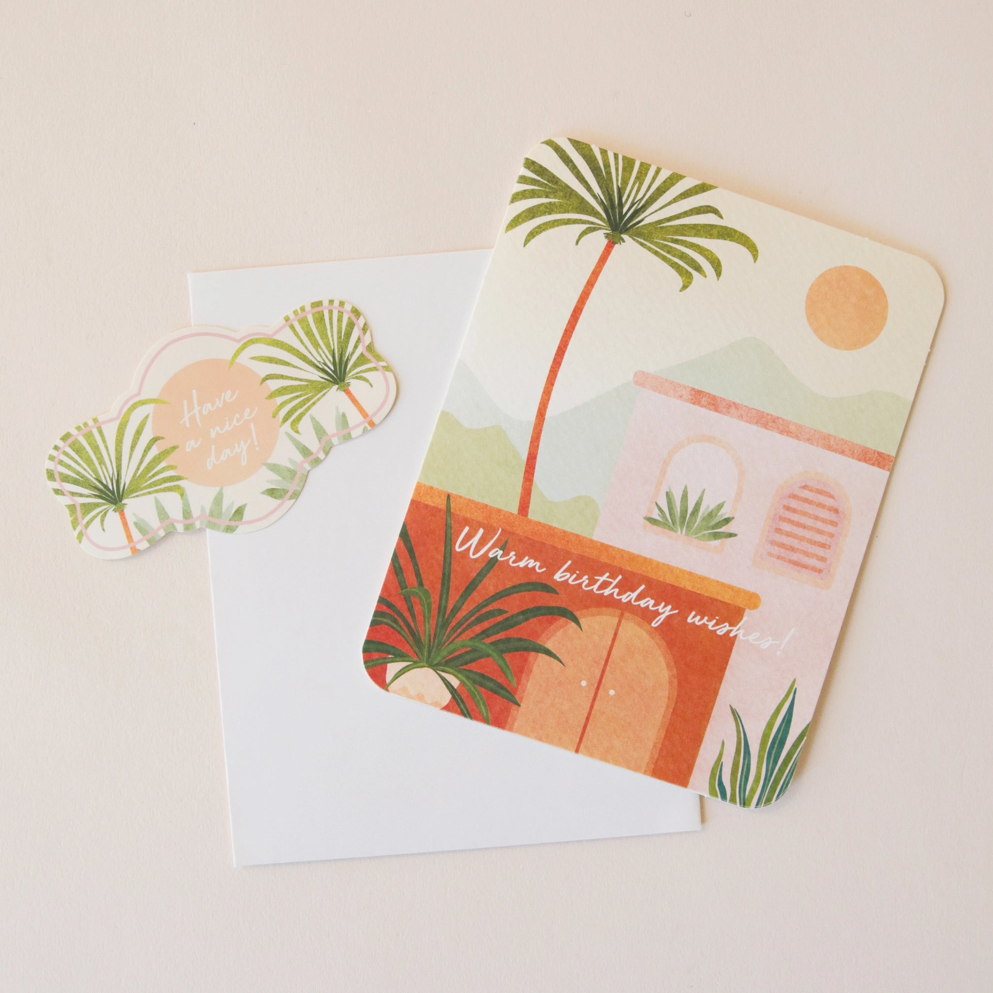 On a neutral background is a card with a sun and palm tree graphic along with a coordinating sticker and text along the front that reads, "Warm birthday wishes".