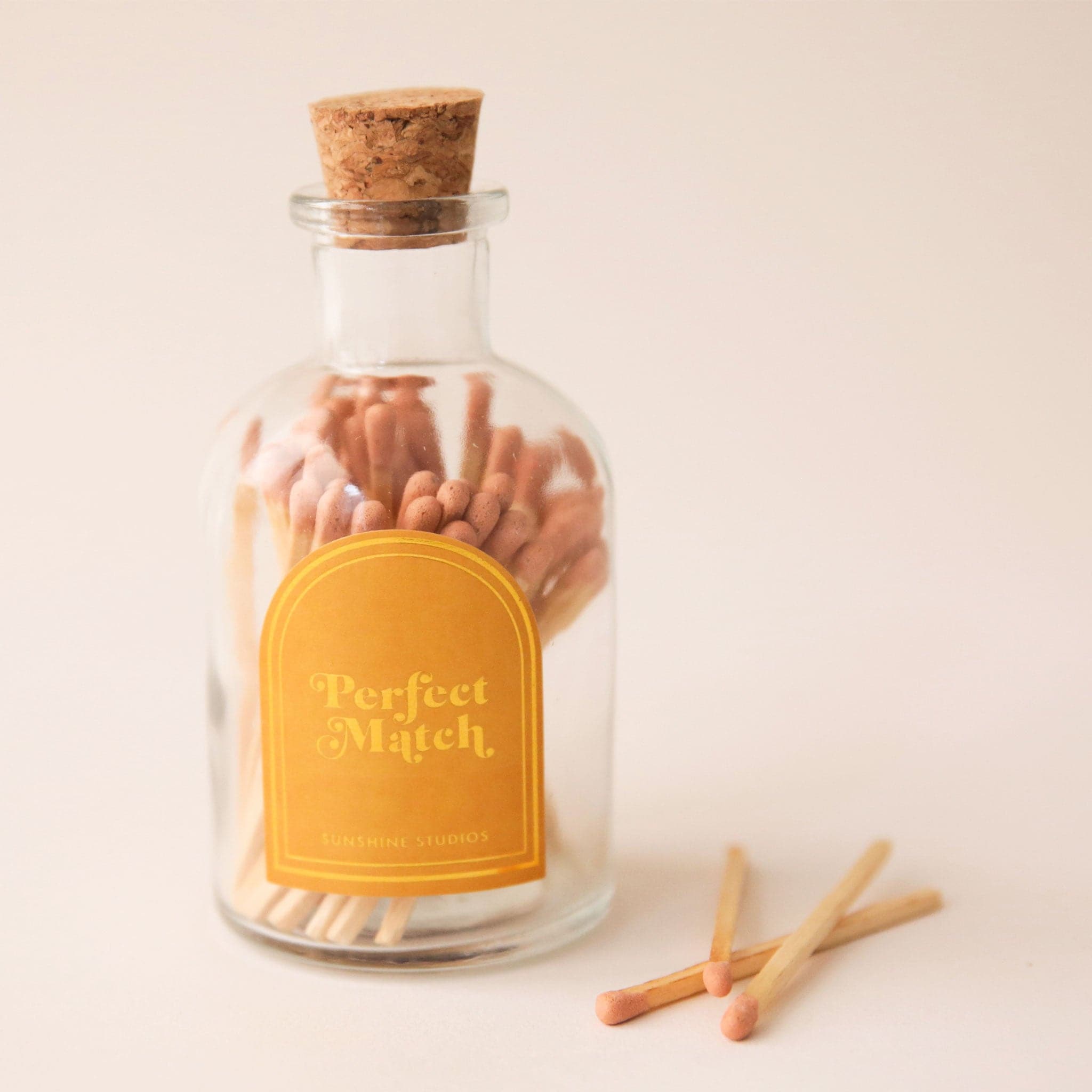 A glass jar of matches with a gold arched label and a cork cap.