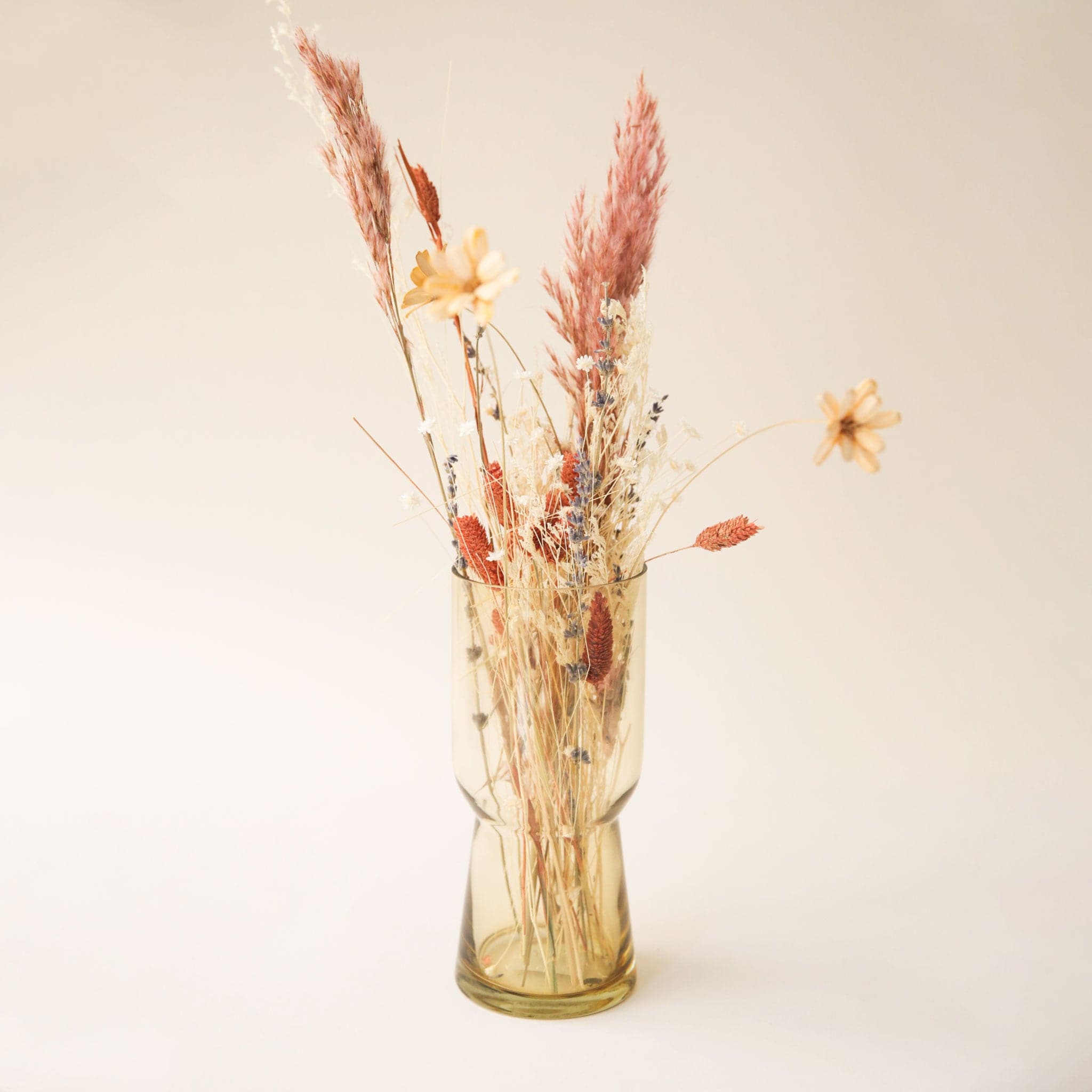 A compote style glass vase in an amber color in front of a white ground with the taller thinner version of the vase filled with dried florals.
