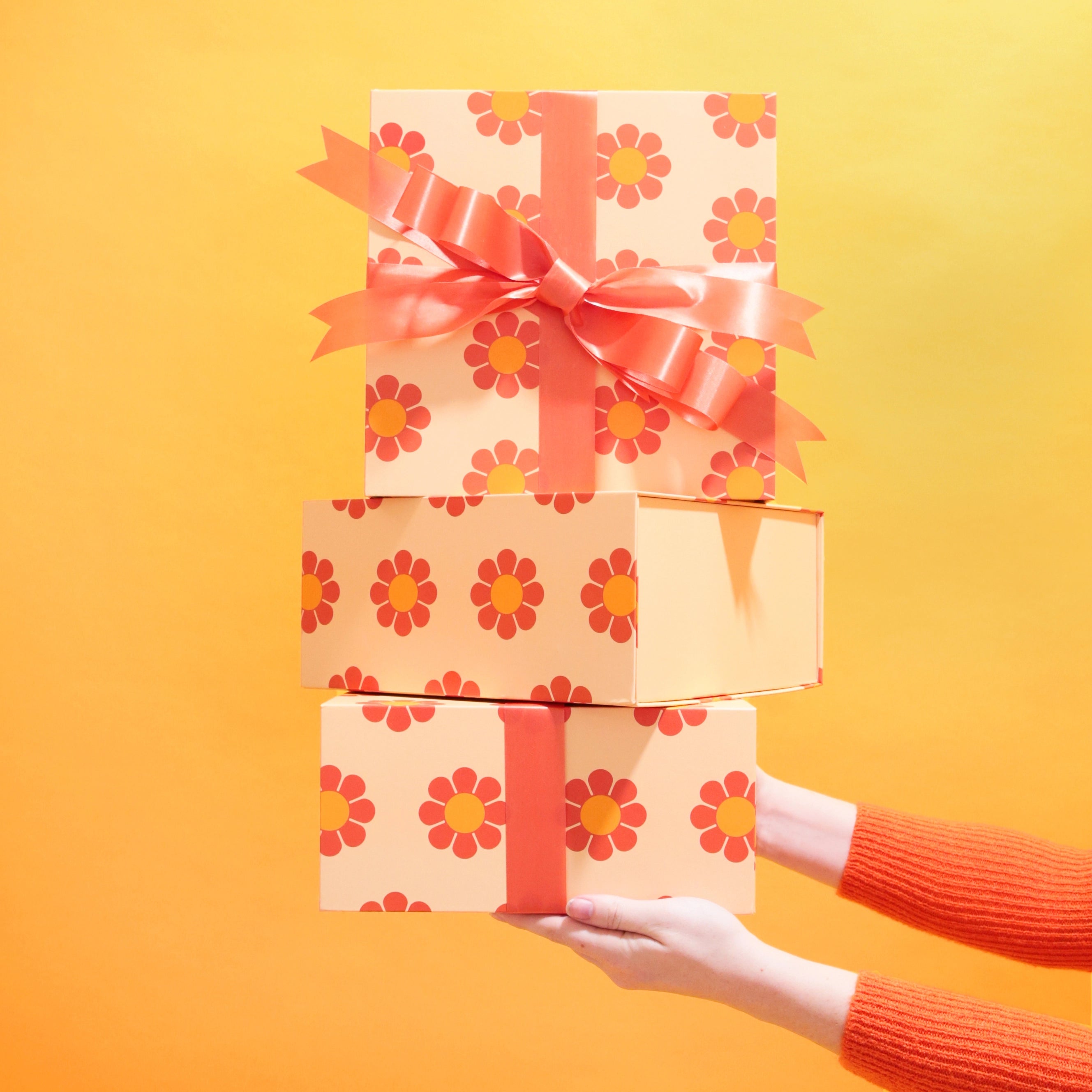 In front of a yellow background is a stack of three orange gift boxes with red and yellow daisies and a red bow not included with purchase.