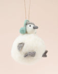 In front of a pink background is a felt penguin ornament. The body is a white, round body. It has two little gray feet and two gray hands. On top is a little white and gray head with a gray nose, two black eyes and green ear muffs.