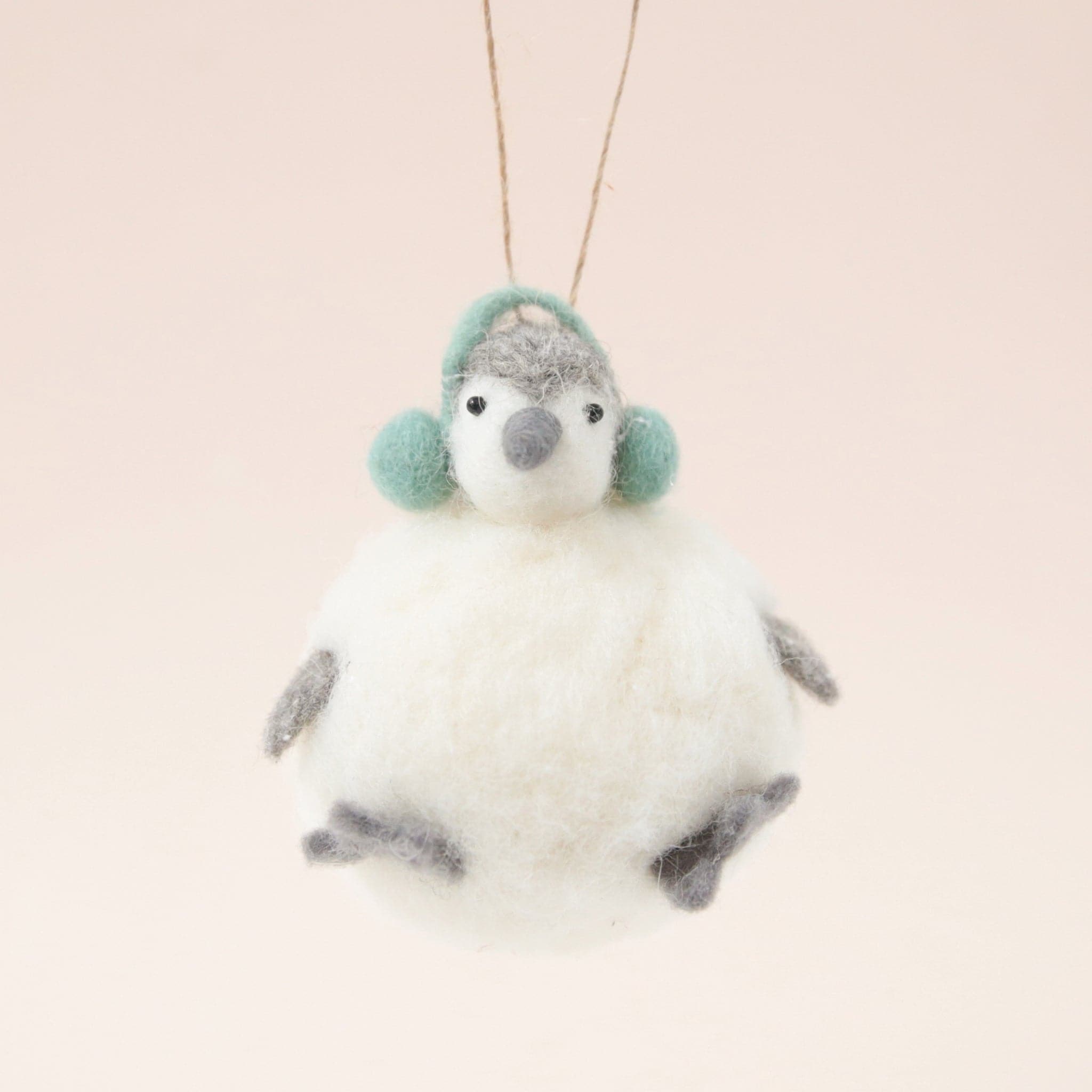 In front of a pink background is a felt penguin ornament. The body is a white, round body. It has two little gray feet and two gray hands. On top is a little white and gray head with a gray nose, two black eyes and green ear muffs.