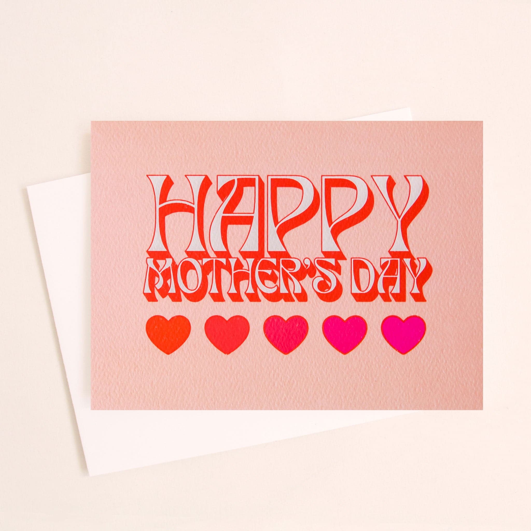 Soft pink card that reads &#39;Happy Mother&#39;s Day&#39; in retro white lettering with a red shadow. Below is a row of five hearts in various reds and pinks. The card is accompanied by a solid white envelope.
