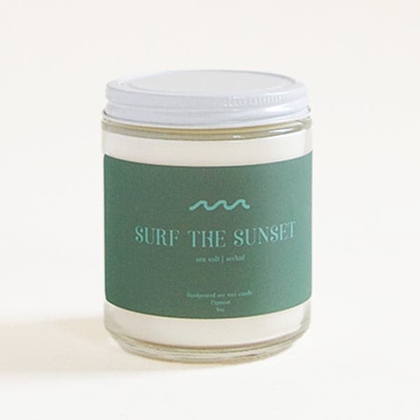 Against a white background sits a clear glass cylinder jar. The jar has a white lid. A dark turquoise sticker wraps around the front of the jar. At the top of the sticker is a turquoise wave. Under is text in the same color that reads ‘surf the sunset.’ 