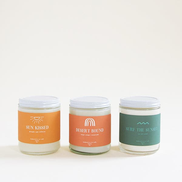 Against a white background sits three clear glass cylinder jars. Each jar has a white lid. The far left jar has a bright orange sticker on the front. It has a white sun at the top and white text that reads ‘sun kissed.’ The middle jar has a terracotta sticker on the front. There is a white rainbow at the top and white text that reads ‘desert bound.’ The last jar has a dark turquoise sticker on the front of the jar. There is a turquoise wave at the top and text that reads ‘surf the sunset.’ 
