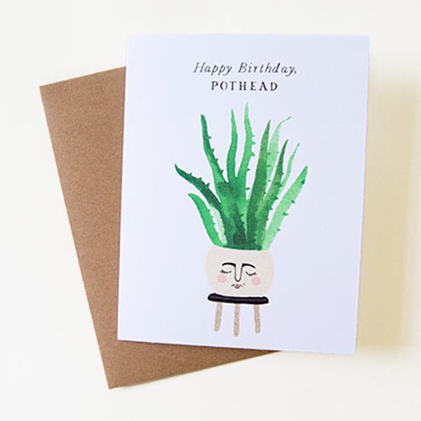On top of a dark brown envelope is a white card. In the middle of the card is a water color painting of a round tan pot sitting on top of a black stool. There is face painted on the front of the pot. Inside the pot is a tall green plant. At the top is black text that reads ‘happy birthday pothead.’ 