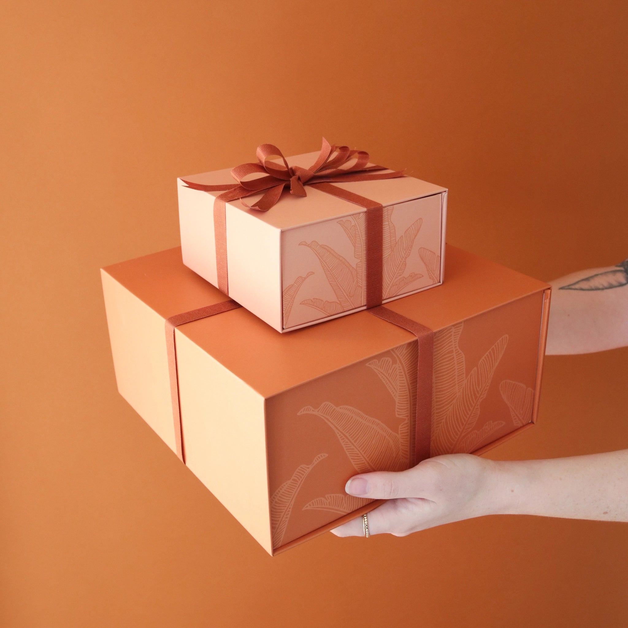A stack of salmon pink gift boxes with a magnetic closure and darker pink bird of paradise line graphic along with a rust colored ribbon tied in a bow, not included with purchase.