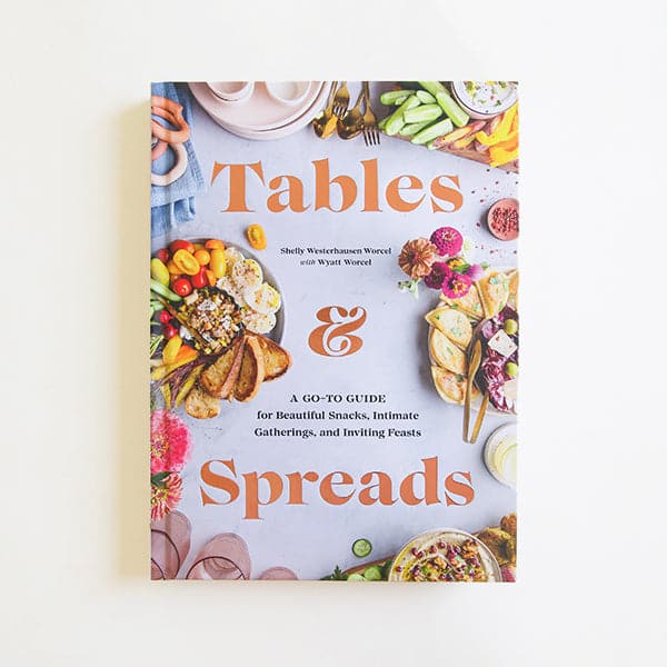 Against a white background is the cover of a cook book. The cover is the birds eye view of a gray table. Around the edges of the cover are trays of food such as vegetables, humus, crackers and flowers. In the middle is rose gold text that reads ‘tables & spreads.’ 