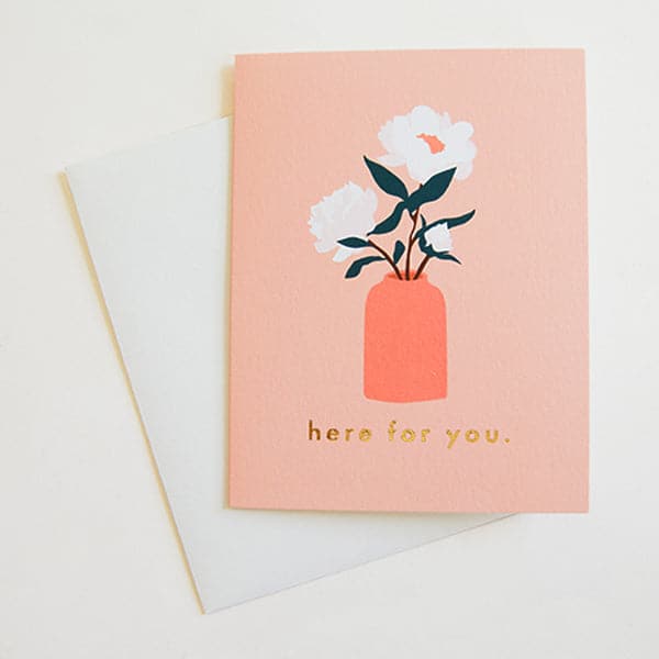 Photo of a peach greeting card on a white background. Greeting card reads "here for you" in lowercase, gold foil letters. There is an illustration of a coral colored vase with 3 white flowers with dark green stems above the writing. Envelope is white. 