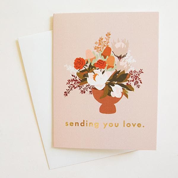 On top of a white envelope is a blush pink card. In the center of the card is a rust tapered flower vase filled with white, pink, red and orange flowers. Below is gold text that reads ‘sending you love.'