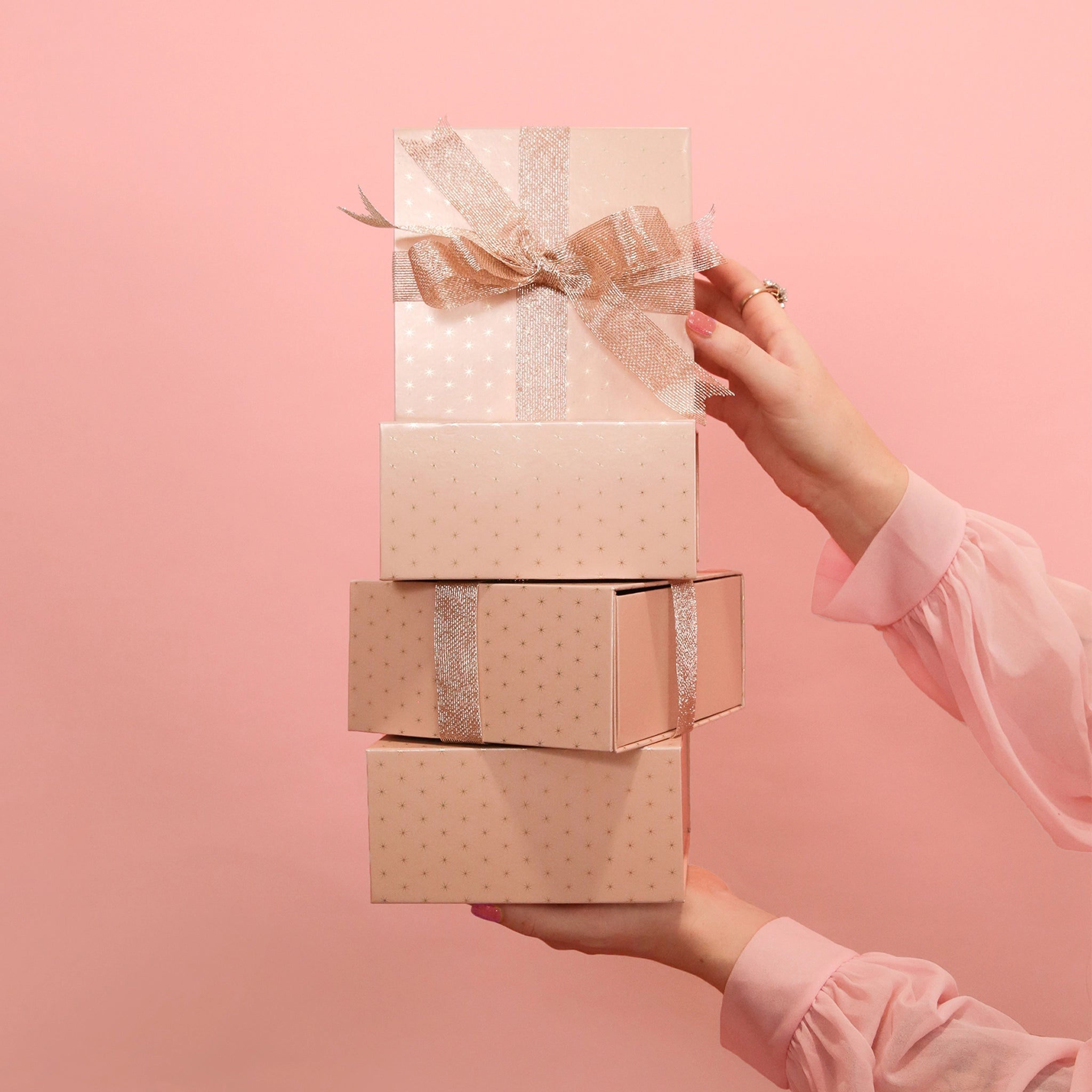In front of a pink background is four light pink gift boxes with a gold foiled star design on four of the sides, the other sides are solid pink. The gift boxes are accented with a sparkle ribbon tied in a bow, not included with purchase.