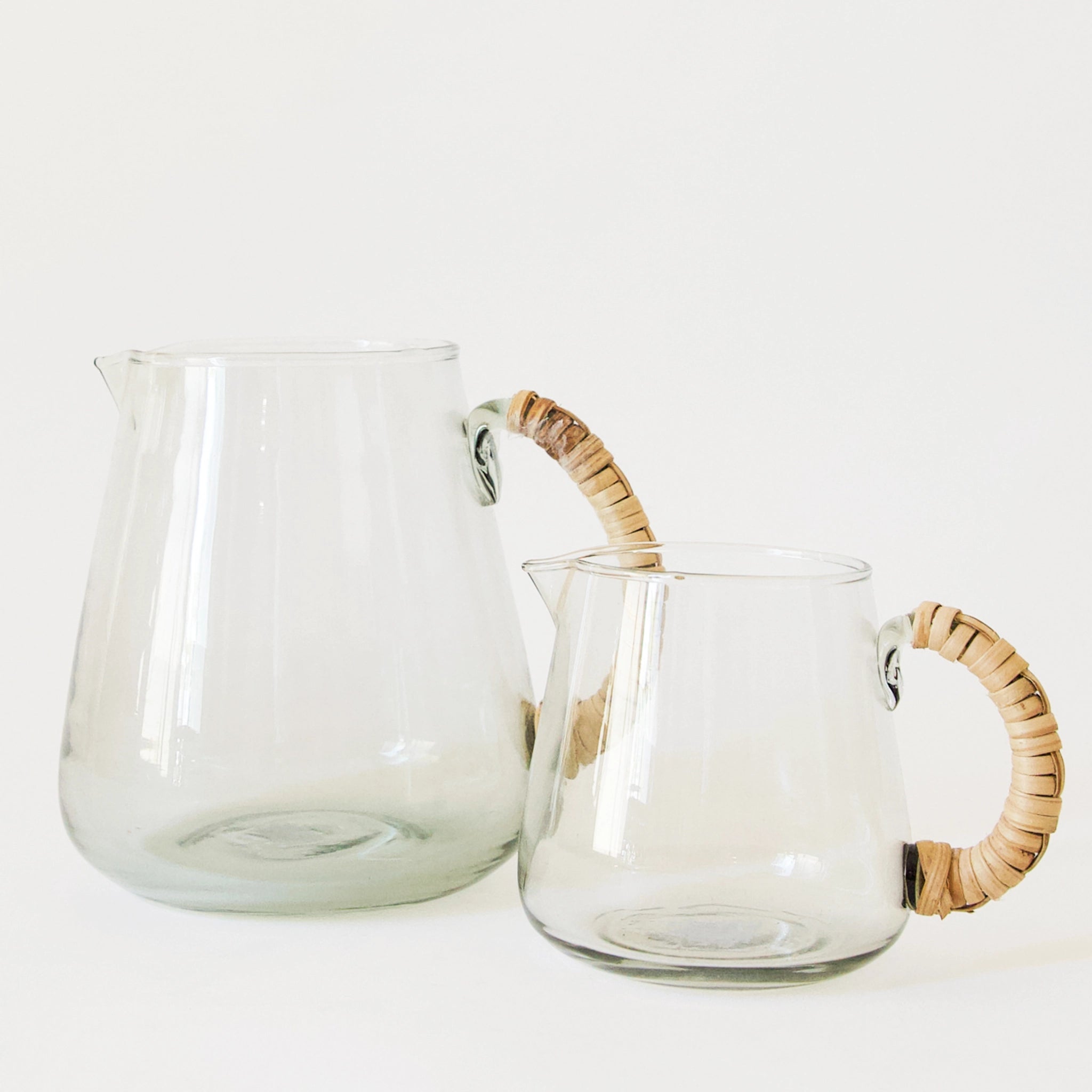 Two glass pitchers, one small and one large, sit side by side. the pitchers have rattan wrapped handles.