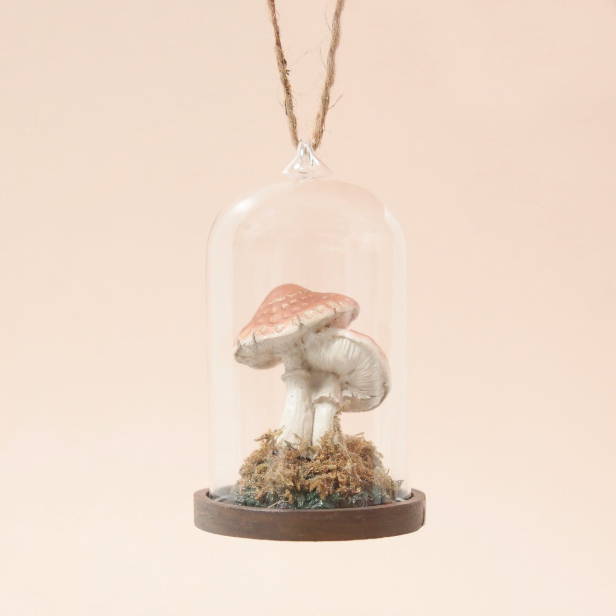 This mushroom ornament features two mushrooms of different sizes with a tan colored top, placed in a glass dome cloche with a wooden base and moss covering the bottom.