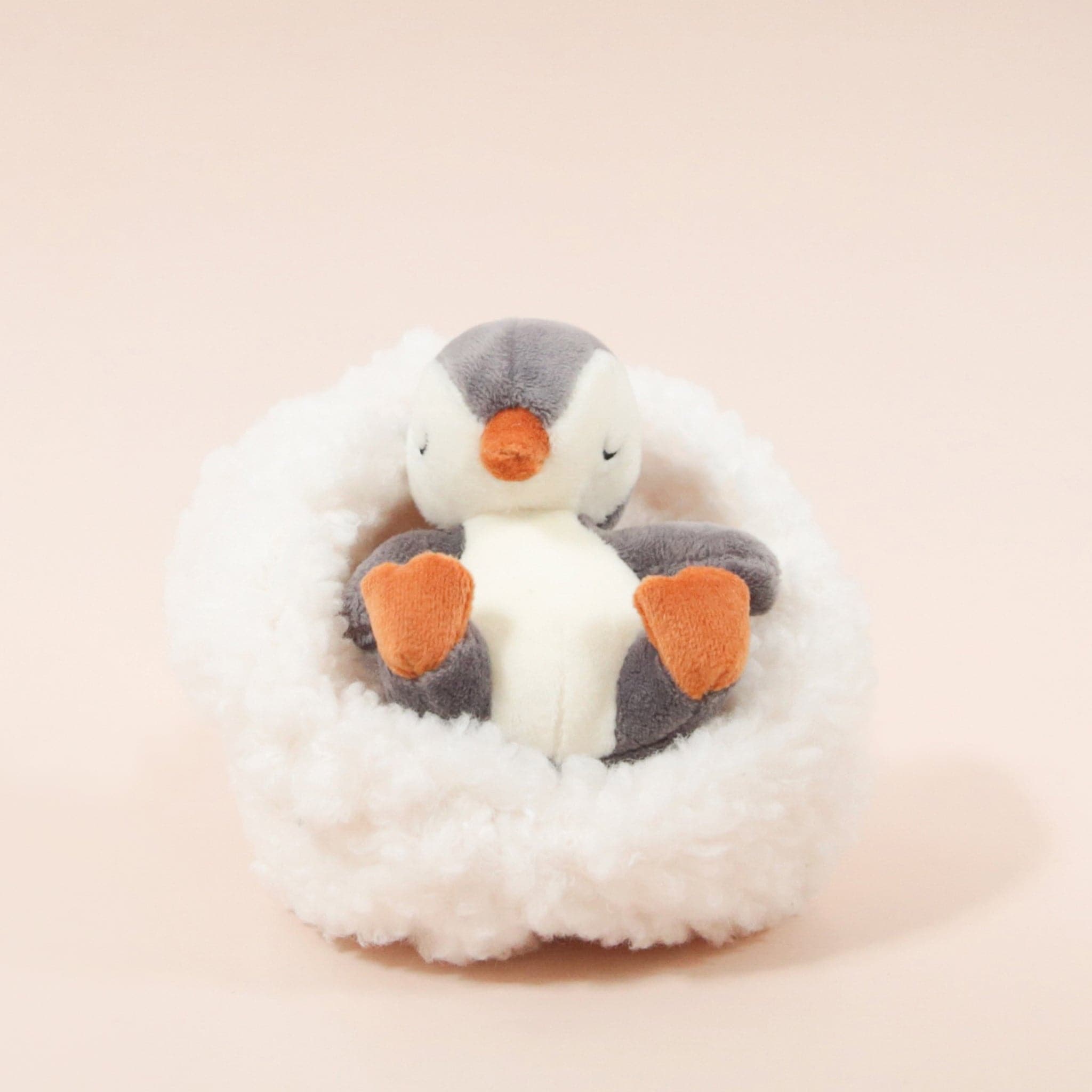 On a cream background is a penguin stuffed animal with a white boucle bed. 