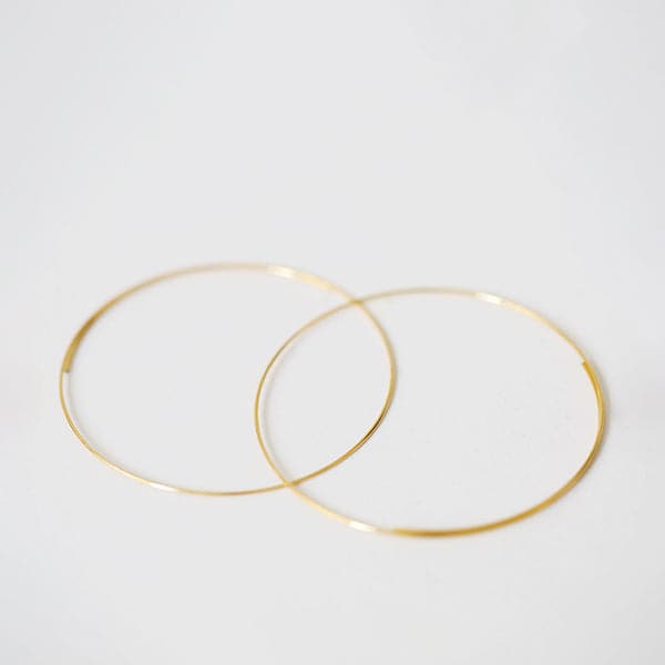 Laying flat on a white background is a pair of thin larger gold hoop earrings.
