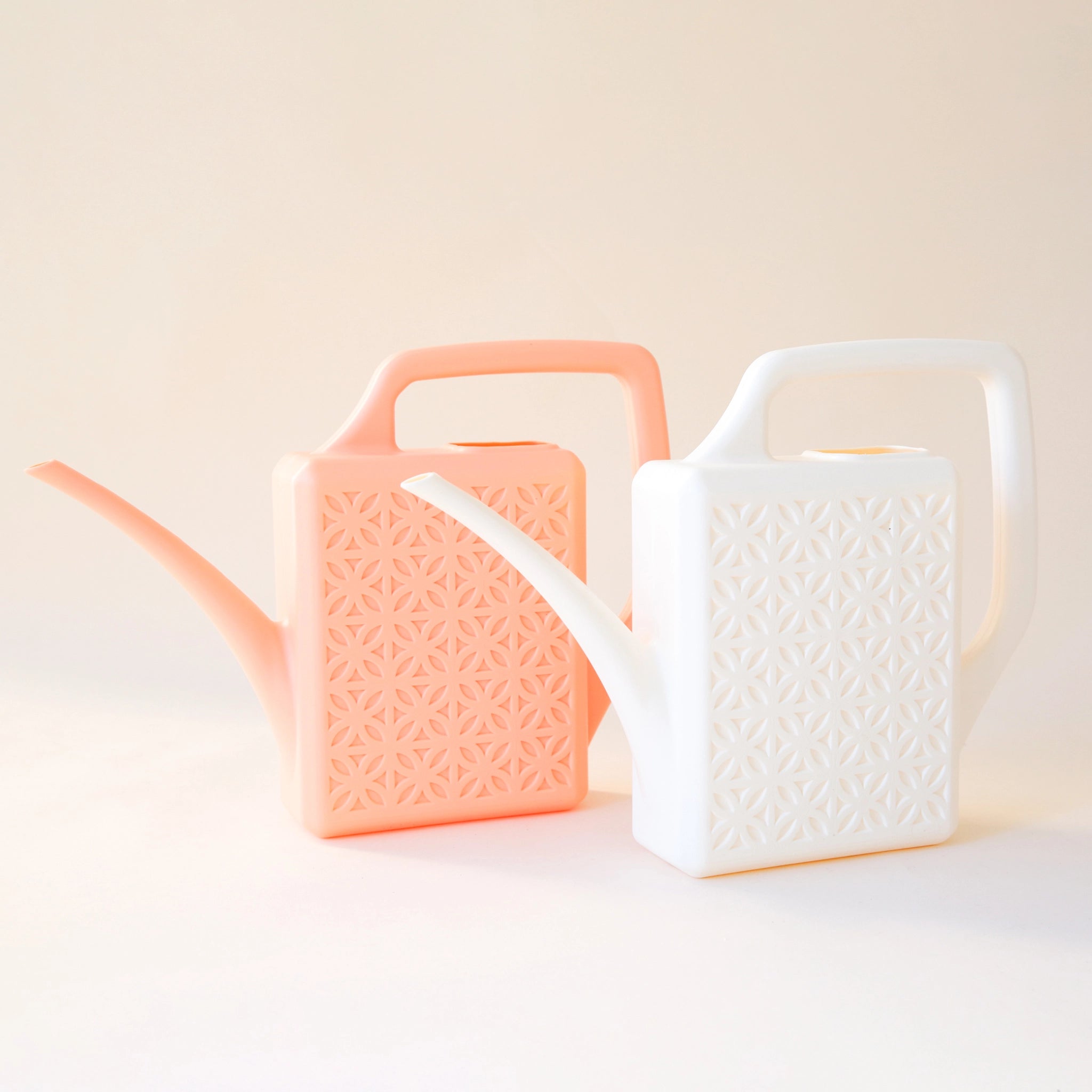 An ivory  and peach plastic watering cans sitting side by side with a narrow spout and square handle and a rectangle breeze block design on the sides.