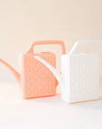 An ivory  and peach plastic watering cans sitting side by side with a narrow spout and square handle and a rectangle breeze block design on the sides.