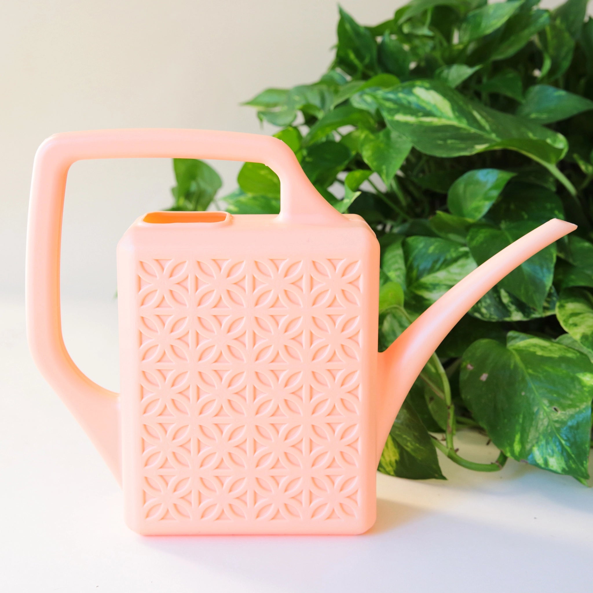 A peach plastic watering can with a narrow spout and square handle and a rectangle breeze block design on the sides.