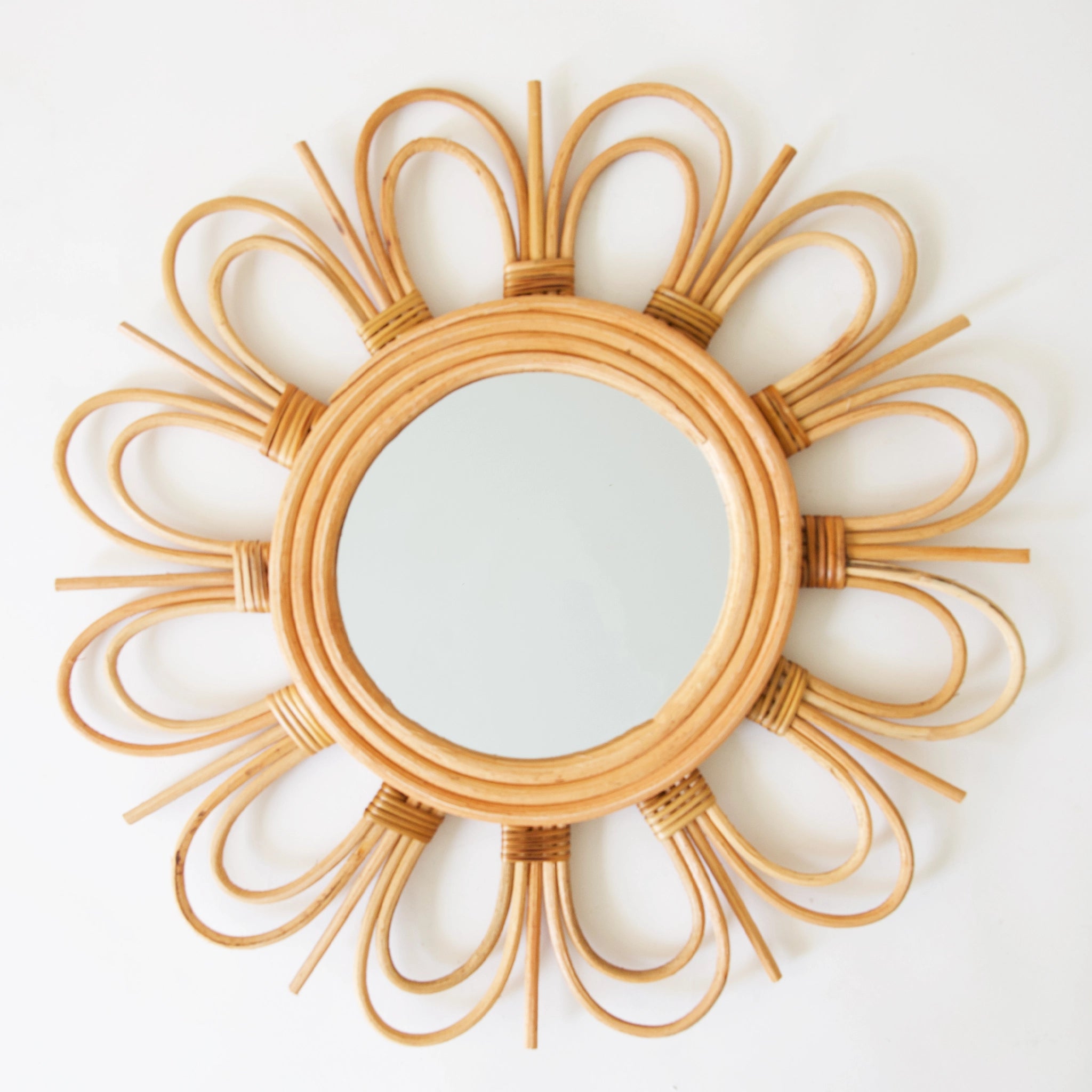 Hanging in front of a white background is a rattan mirror. The mirror is round with three layers of rattan surrounding the outside of the mirror. Around the last layer is a row of small arched with a larger arch over. In between the arches is a straight piece of rattan.