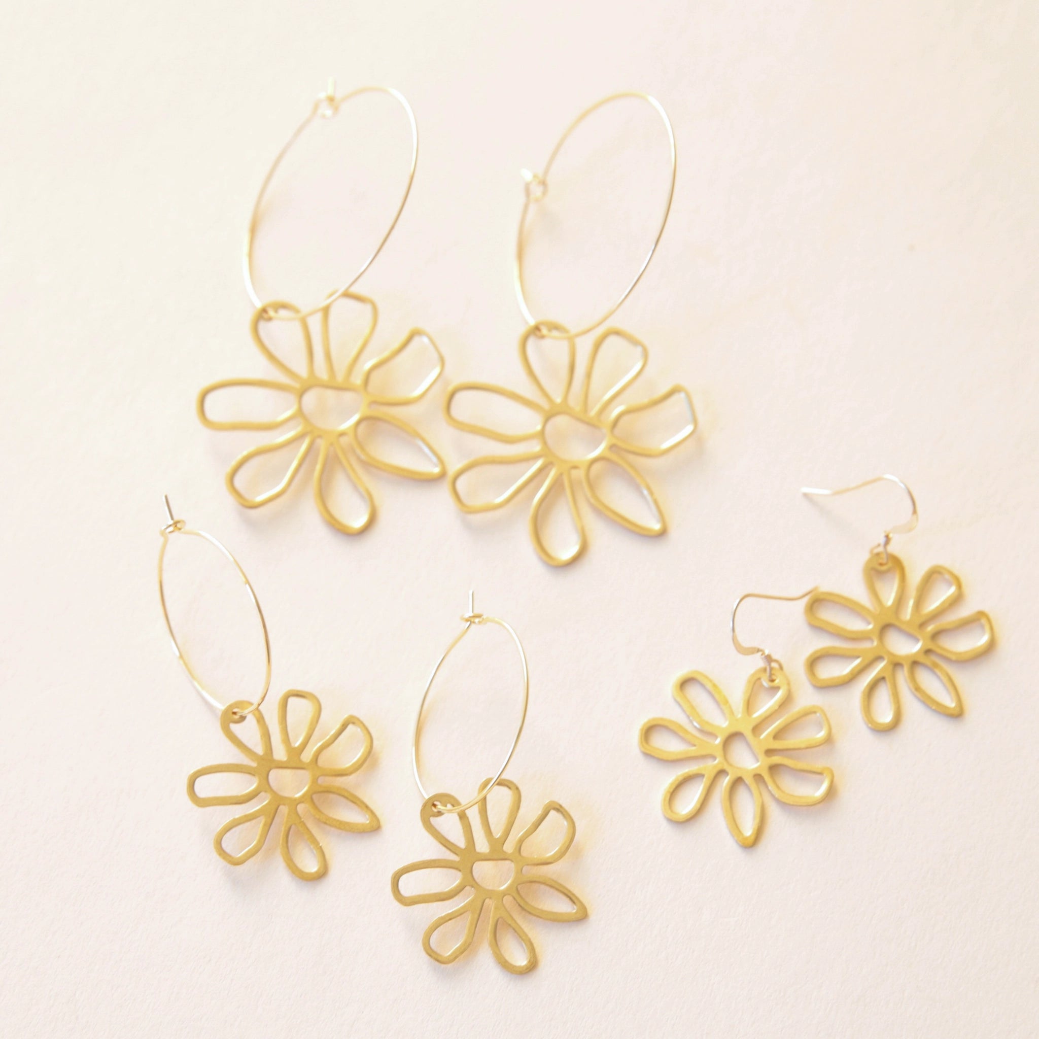 On a cream background is three different pairs of earrings, two different sized pairs of hoops and gold flower charms hanging off the bottom and a pair that has a gold hook earring with the same gold flower charm attached to that. 