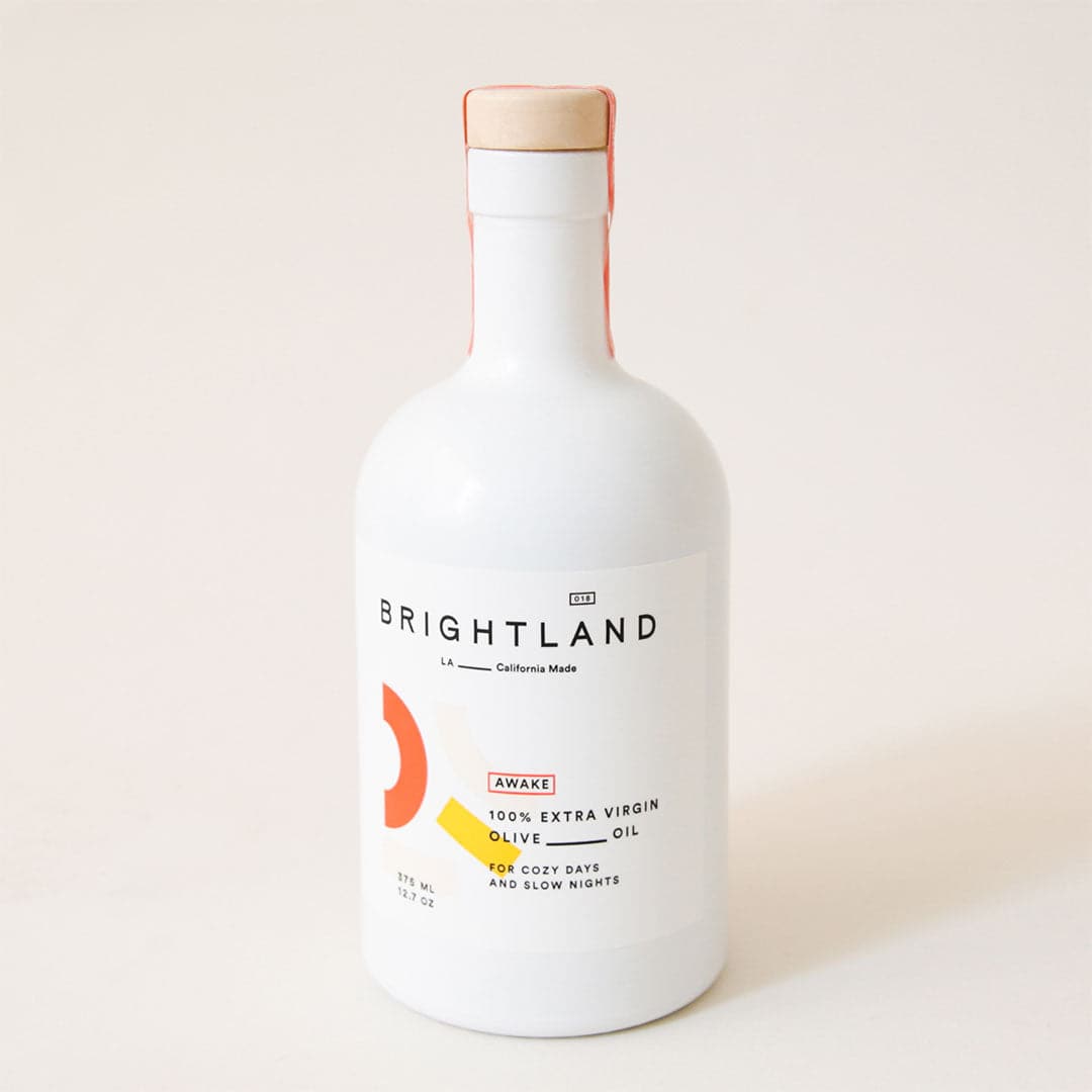 A white opaque bottle with light brown stopper and label with text &quot;Brightland. LA California Made. Awake. 100% Extra Virgin Olive Oil. For cozy days and slow nights.&quot; 