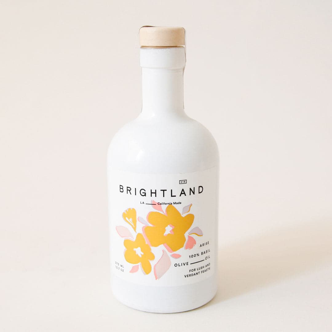 White opaque bottle with light brown stopper. White label says &quot;Brightland. LA, California Made. 100% Basil Olive Oil. For Lush and Verdant Feasts,&quot; with a watercolor illustration of yellow and pink flowers.