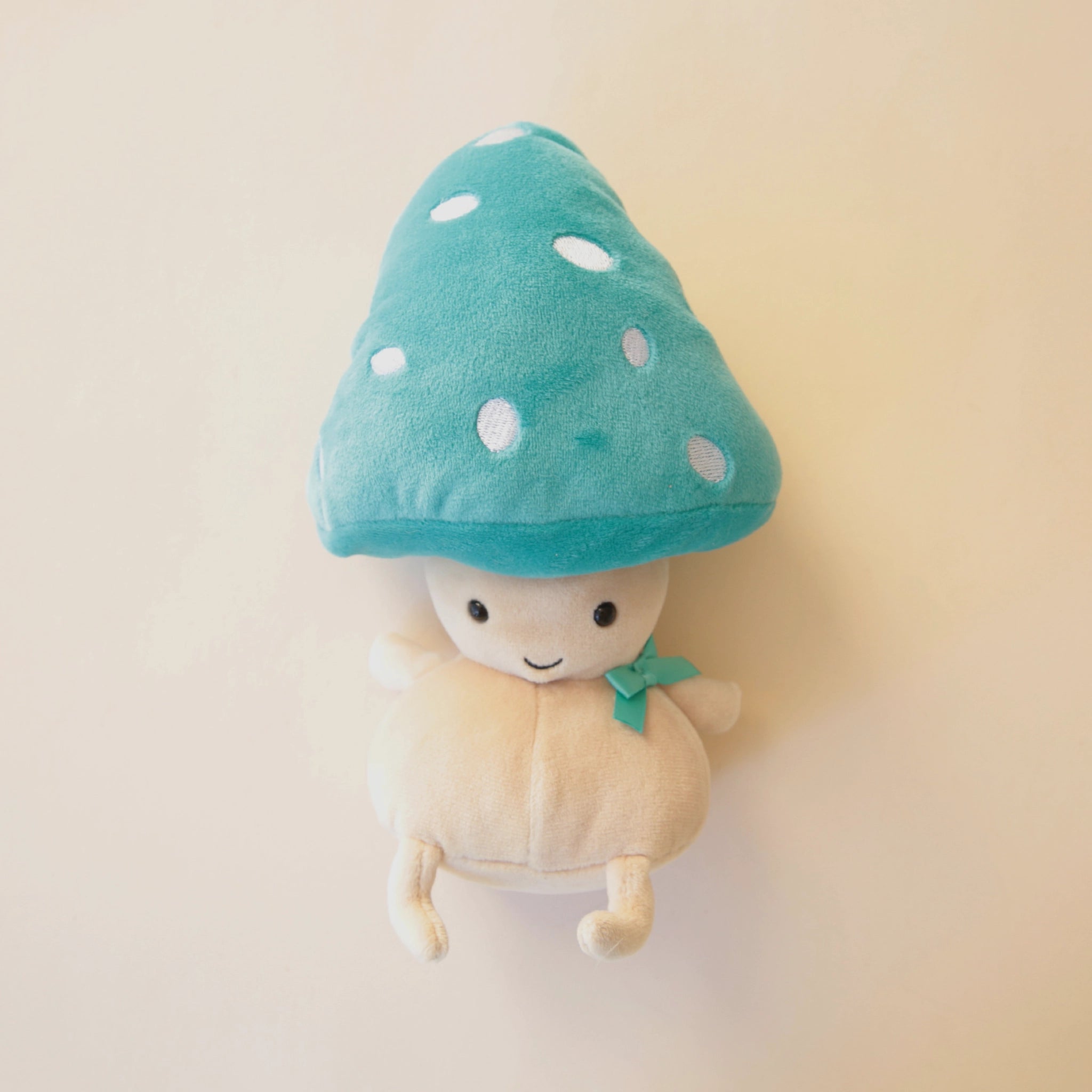 A oatmeal colored mushroom character with black eyes and a small smiling mouth along with a turquoise bowtie and the same colored mushroom hat with white spots.