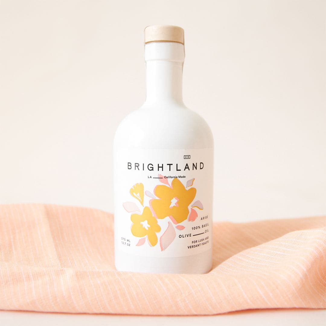 White opaque bottle sits on a pink with white stripes tea towel, with light brown stopper. White label says &quot;Brightland. LA, California Made. 100% Basil Olive Oil. For Lush and Verdant Feasts,&quot; with a watercolor illustration of yellow and pink flowers.