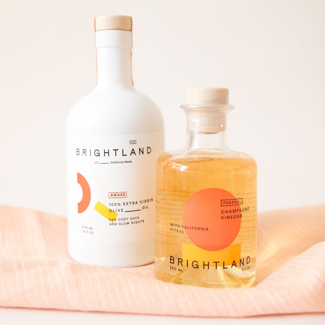 Two bottles side to side on a pale pink tea towel. The left is a white opaque bottle with light brown stopper and label with text &quot;Brightland. LA California Made. Awake. 100% Extra Virgin Olive Oil. For cozy days and slow nights.&quot; The right is a clear bottle with a golden liquid and light brown stopper, with clear label with text &quot;Brightland. With California Citrus. Champagne vinegar.&quot;