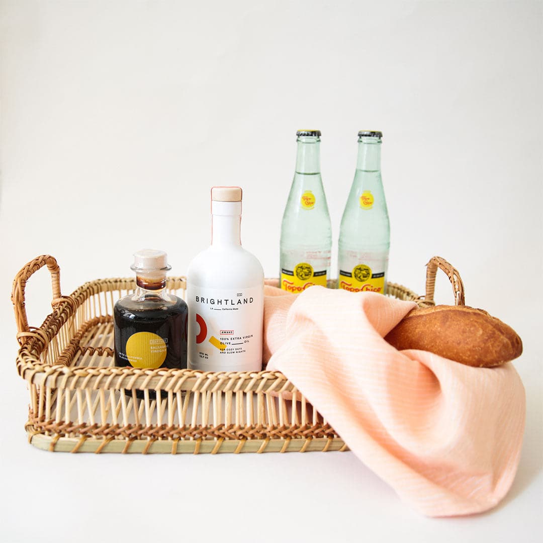 A rattan tray with a bottle of Brightland balsamic vinegar, olive oil, two bottles of Topo Chico sparkling water, a baguette loaf and pale pink tea towel.