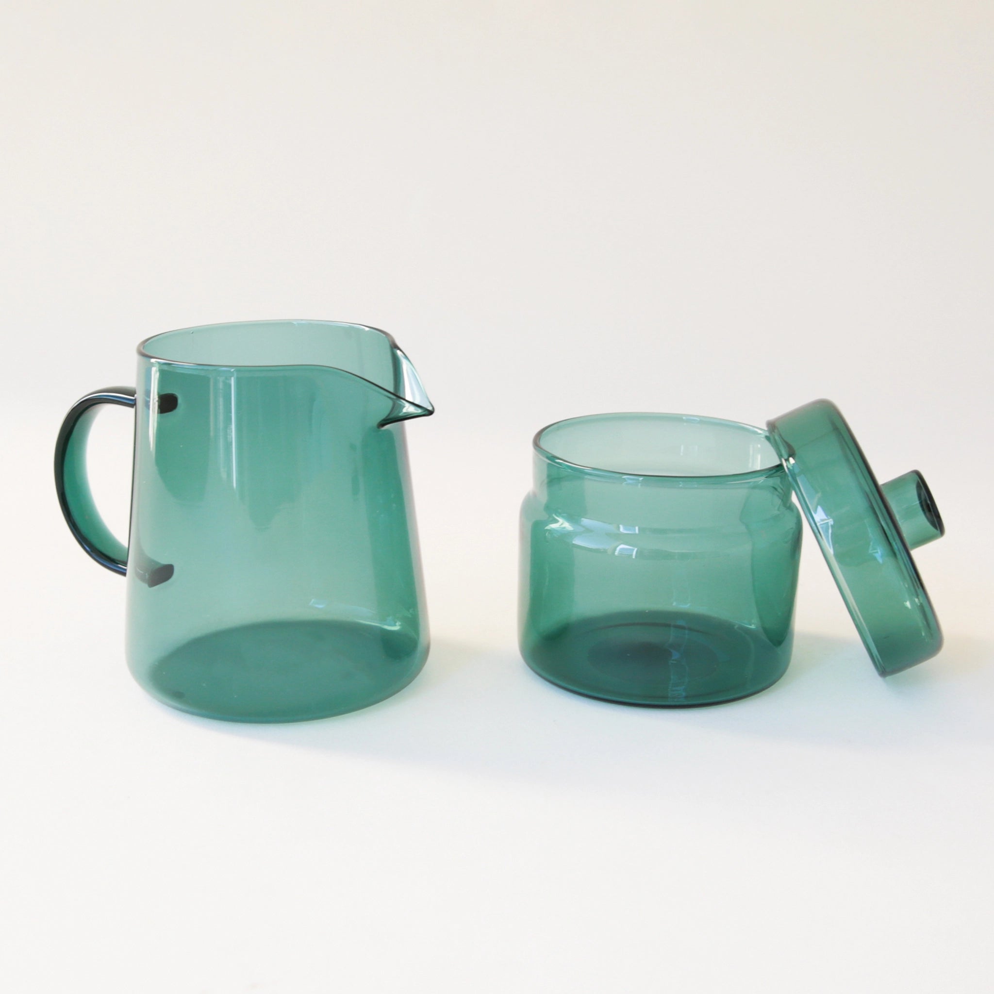 A round glass teal jar with a coordinating glass lid and a round but flat knob on top photographed here besides a matching teal pitcher.