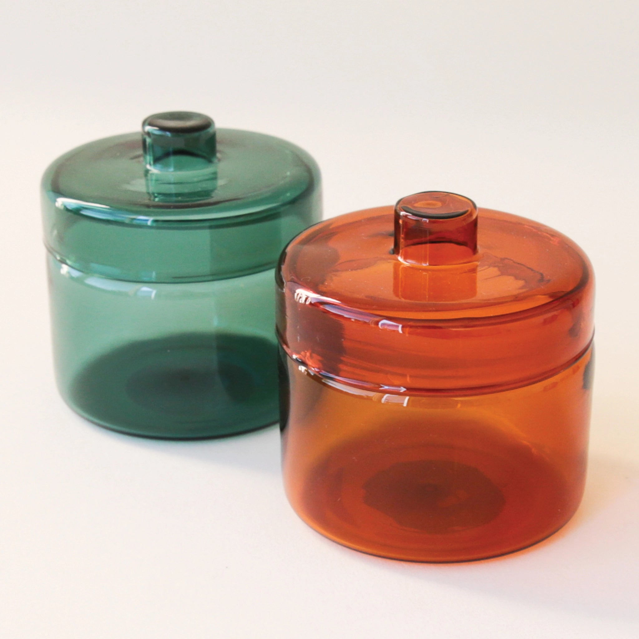 A round glass teal jar with a coordinating glass lid and a round but flat knob on top photographed here next to the same glass jar in a different burnt orange color way.