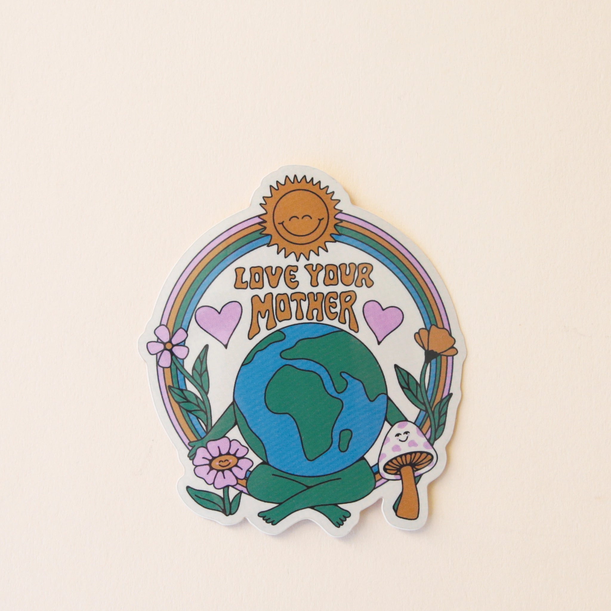 A round vinyl sticker with the planet earth in the center with green legs and arms along with a rainbow above it and a smiling sun in the center of that. Next to the earth is daisy, heart and mushroom graphics.