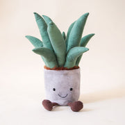 A stuffed animal aloe vera plant with a grey pot, two brown legs and a light green spiky aloe leaves along with a smiling face on the grey planter area.