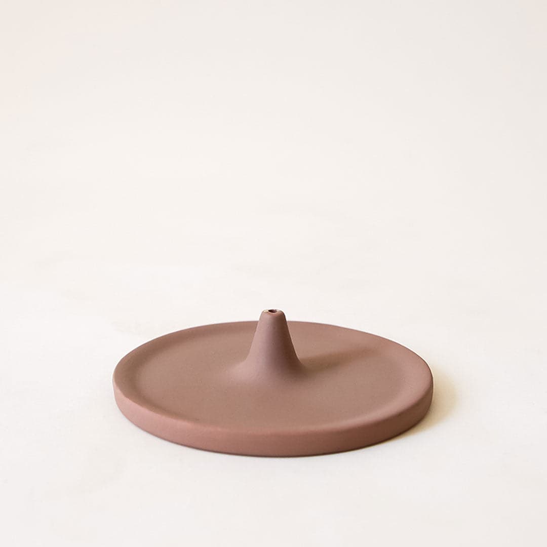 A brown incense holder that has a circular shape with a point in the center that is raised with a hole to place your incense in. 