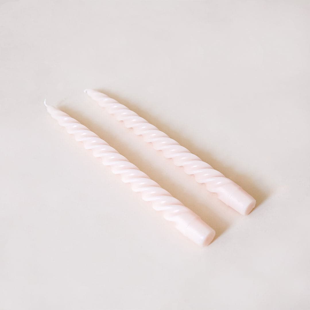 Pair of two blush pink, twisted taper candles with white cotton wicks lay beside each other against a white background.